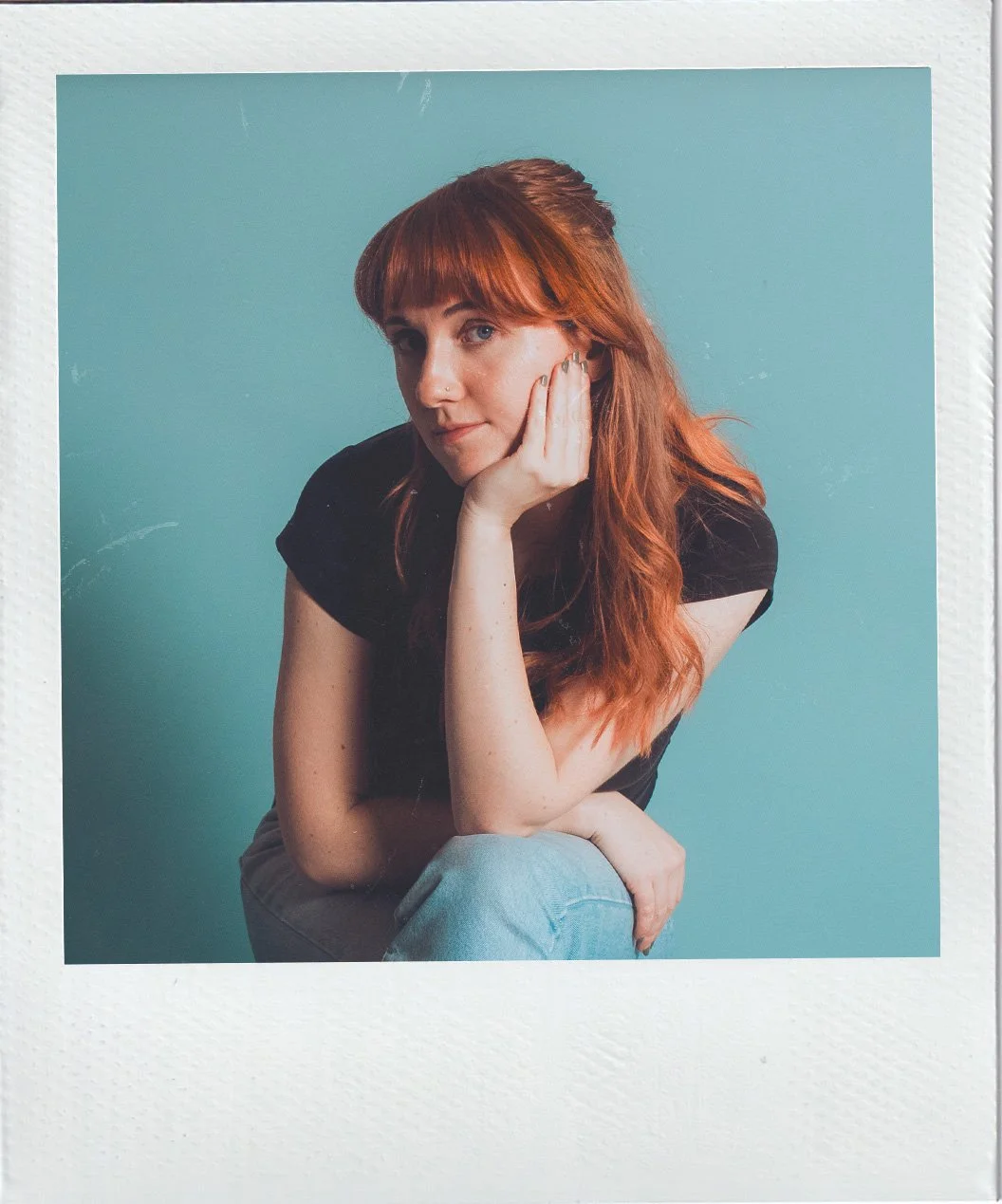 A woman with red hair, sitting against a teal background, resting her chin on her hand, wearing a black shirt and blue jeans.
