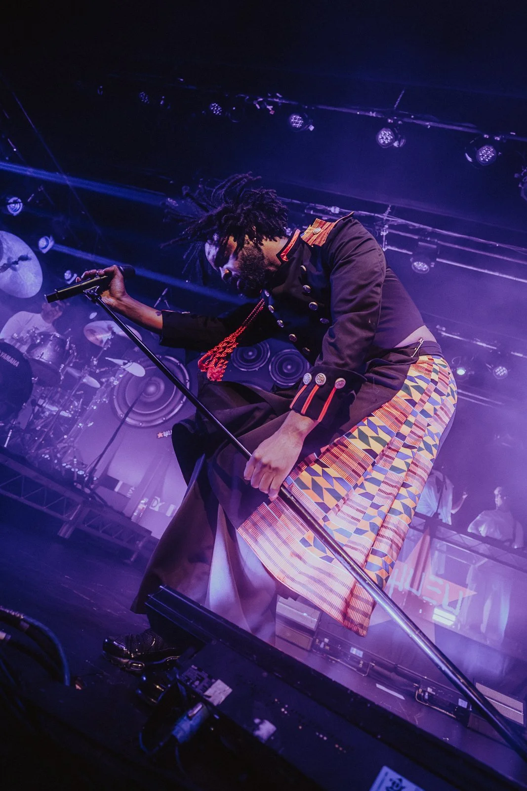 Musician kneeling on stage, holding a microphone, with drum set and DJ equipment in background, under purple stage lights.