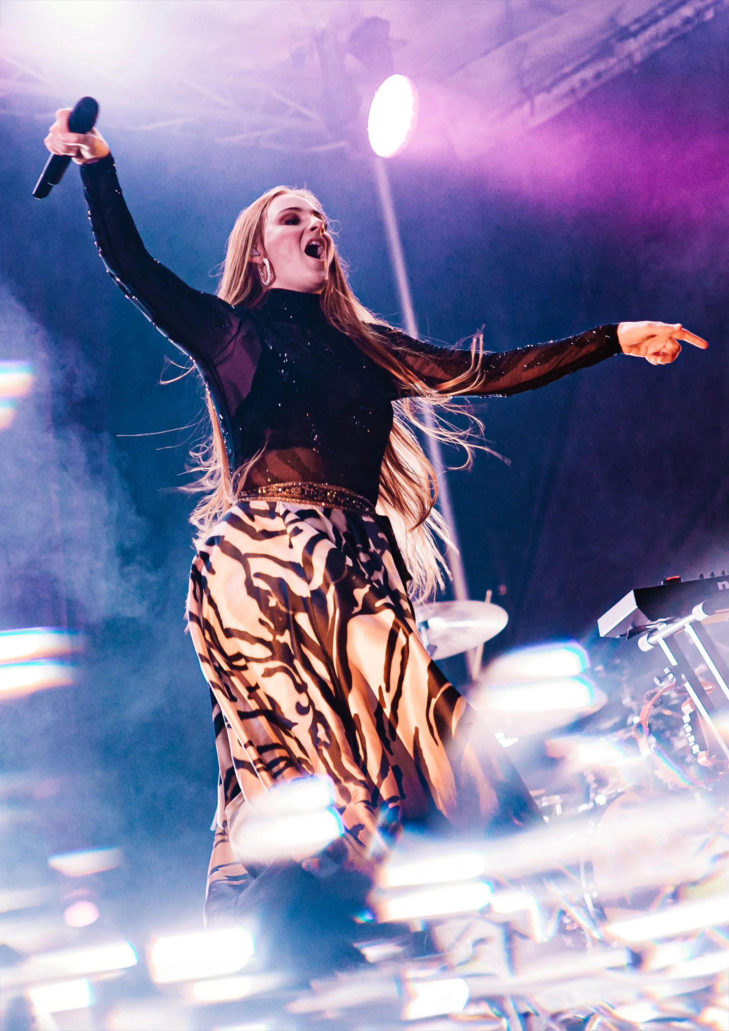 A woman singing into a microphone on stage with colorful lighting and electronic music equipment around her.