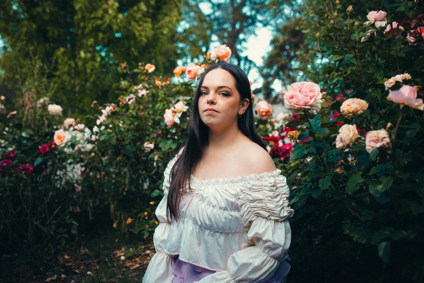 A woman with long dark hair wearing an off-shoulder white blouse stands in a rose garden.