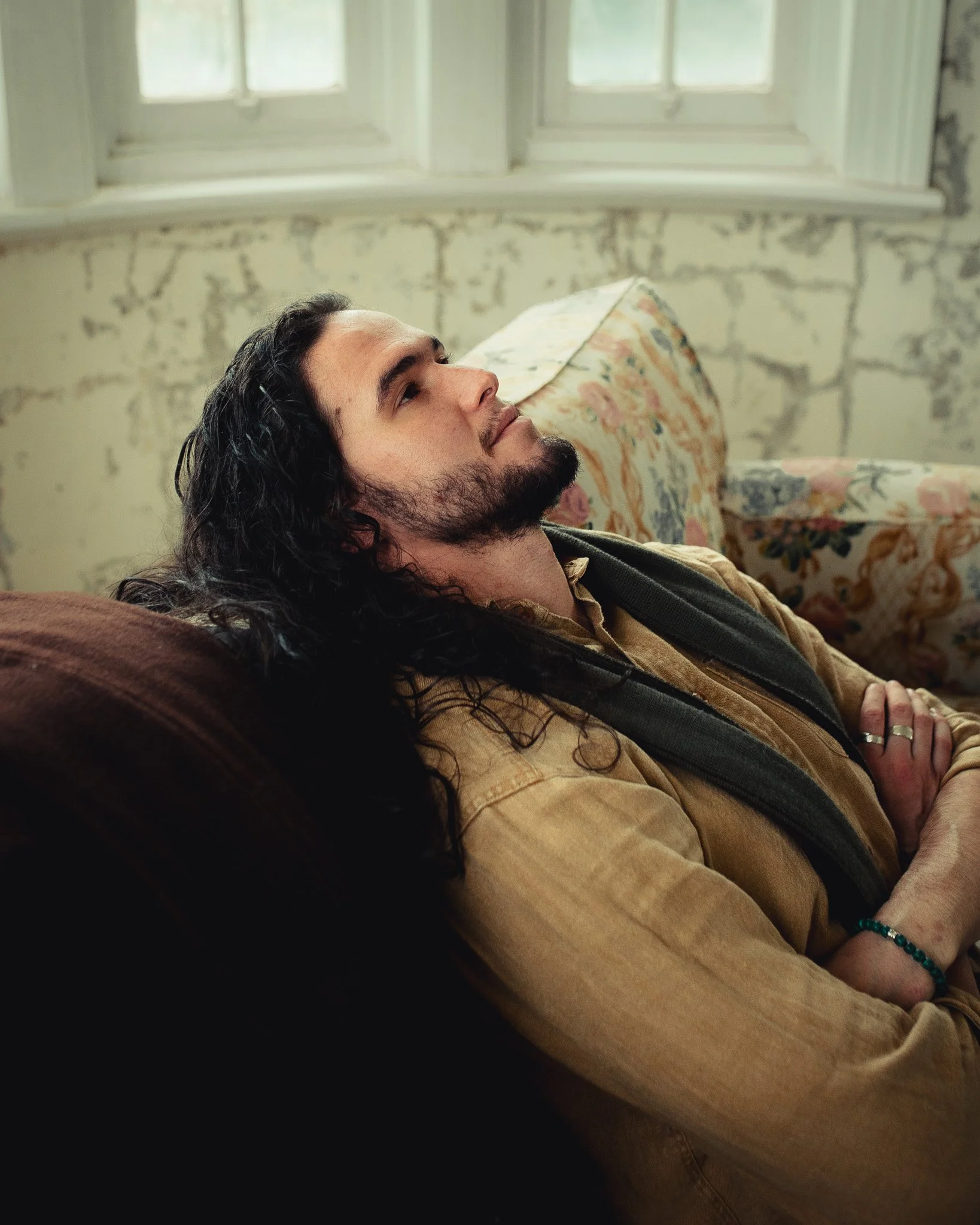 A man with long dark curly hair, a beard, and a mustache is lying on a sofa with his head propped on a floral cushion. He has closed eyes and a relaxed expression, wearing a tan jacket and a black shirt. Behind him is a wall with peeling paint and tw