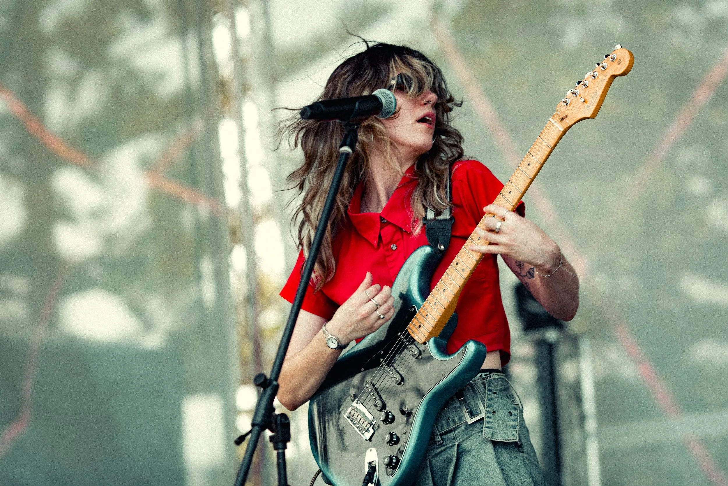A female musician singing into a microphone and playing an electric guitar on stage, wearing a red shirt and jeans