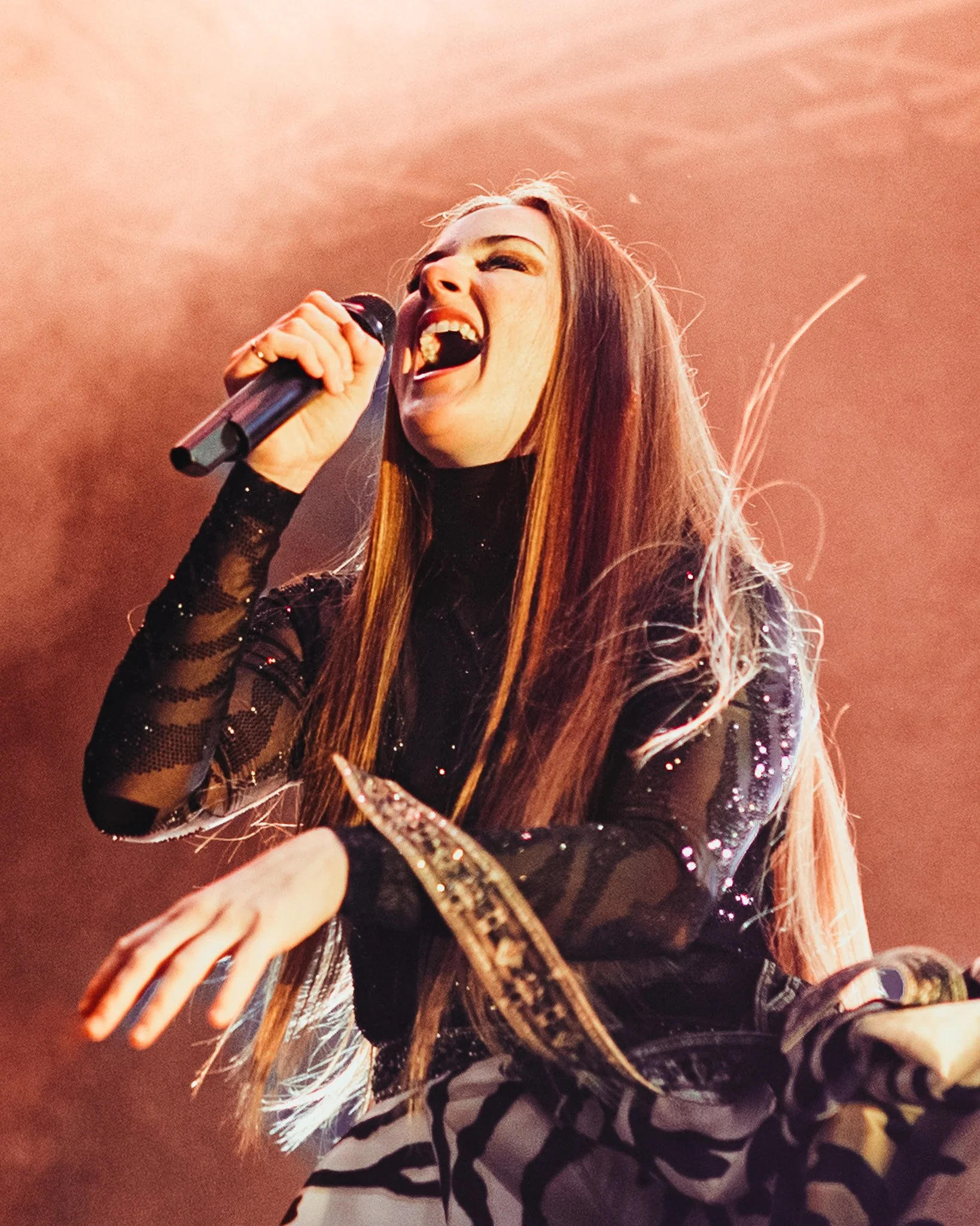 A woman singing passionately into a microphone on stage, with long hair and wearing a black and white animal print outfit.