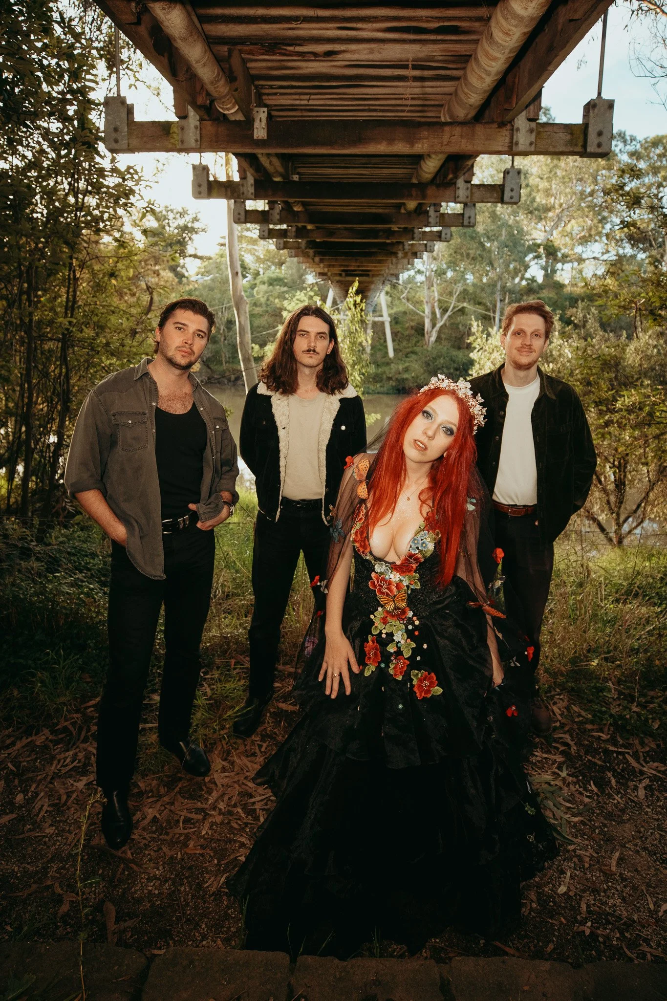 A group of five people, four men and one woman, standing outdoors under a bridge or platform with trees and a lake in the background. The woman in front has long red hair, is wearing a black dress with floral designs, and a floral headpiece. The men 
