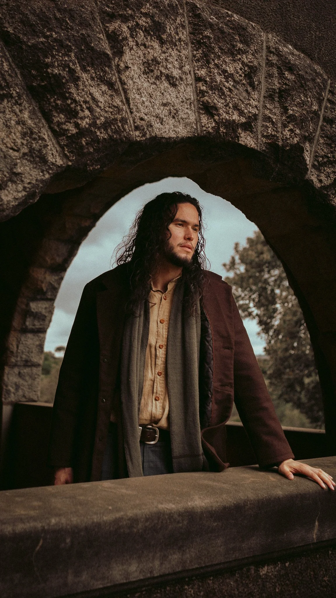 A man with long dark hair standing under a stone archway, looking contemplative and resting his hand on a stone surface.