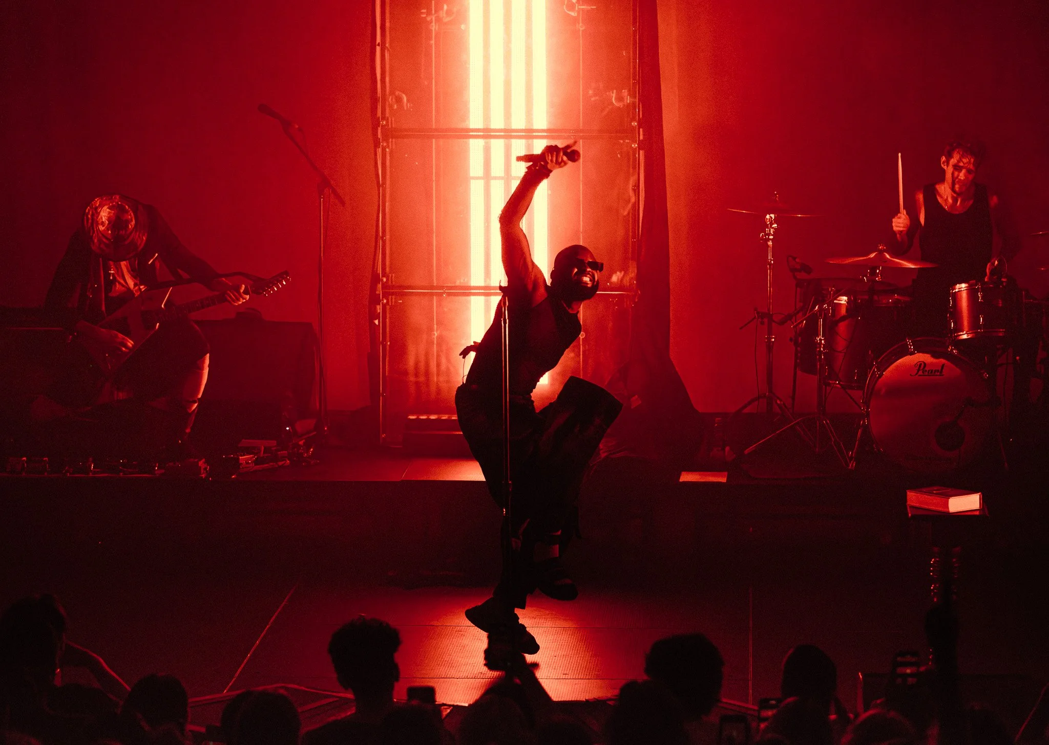 A band performing on stage illuminated by red lighting, with three musicians visible: a guitarist on the left, a lead singer kneeling at the center holding a microphone, and a drummer on the right.