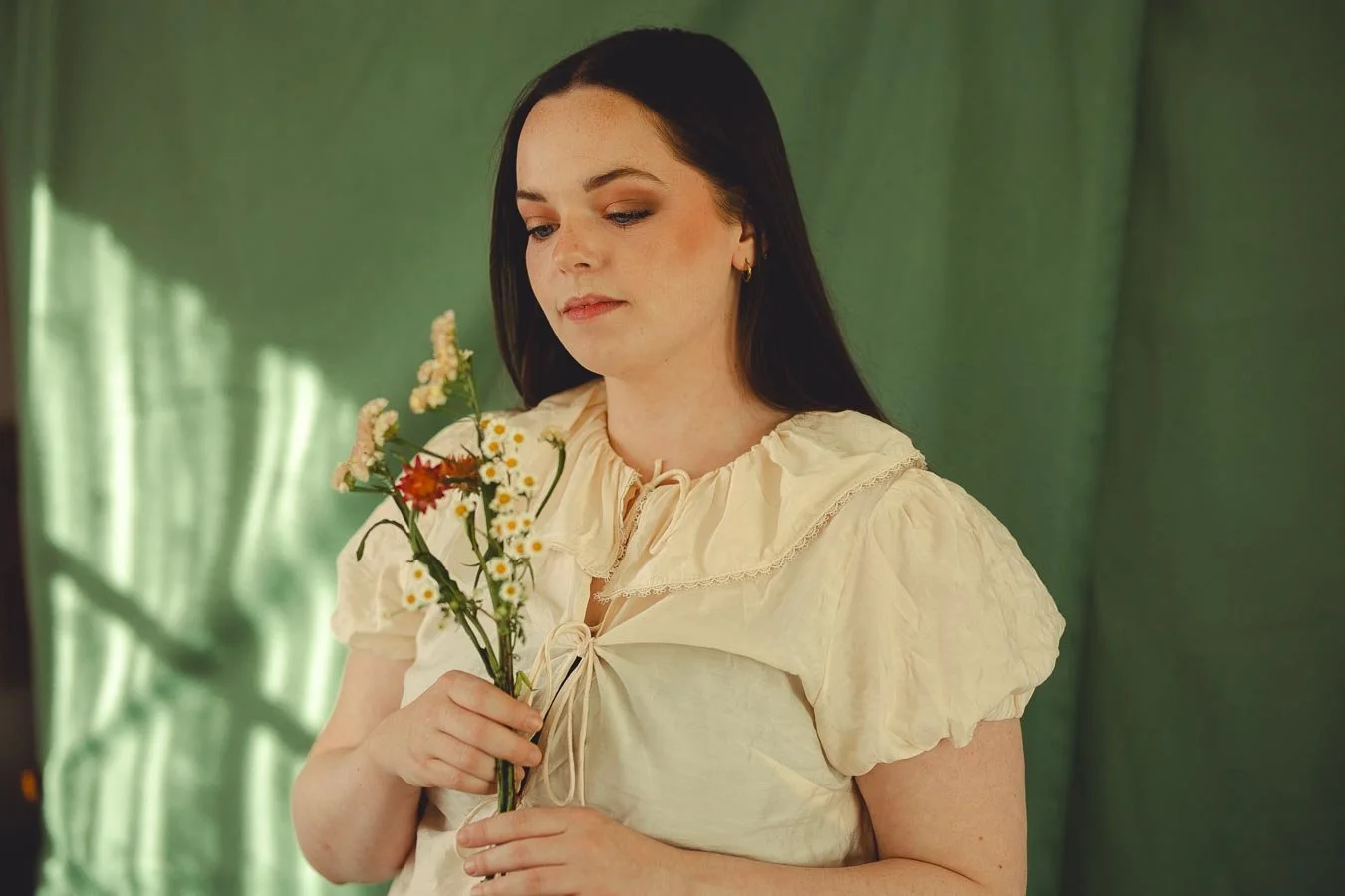 A woman with dark hair holding a small bouquet of wildflowers, standing against a green background.