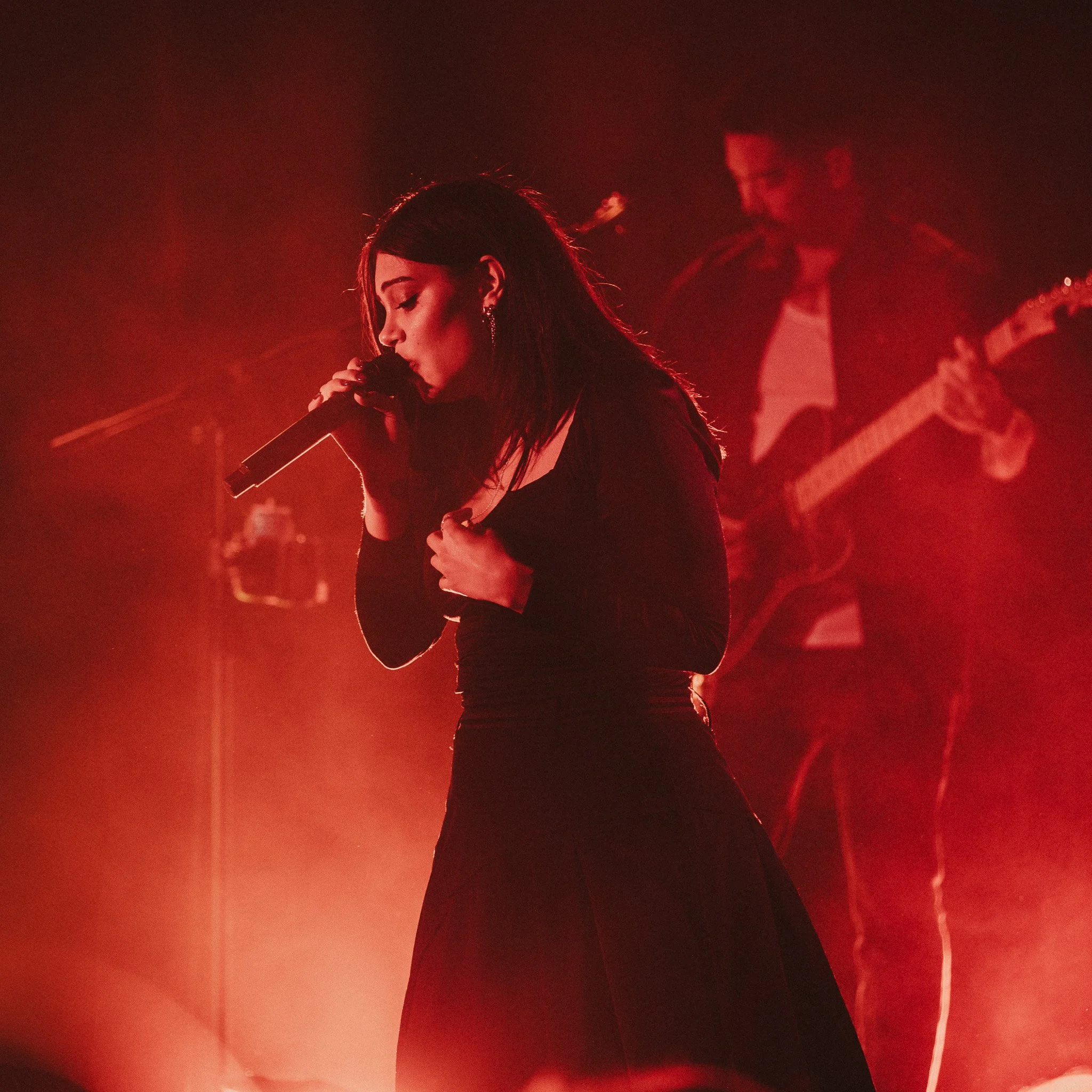 A woman singing into a microphone with a guitarist in the background, all illuminated by red stage lighting.