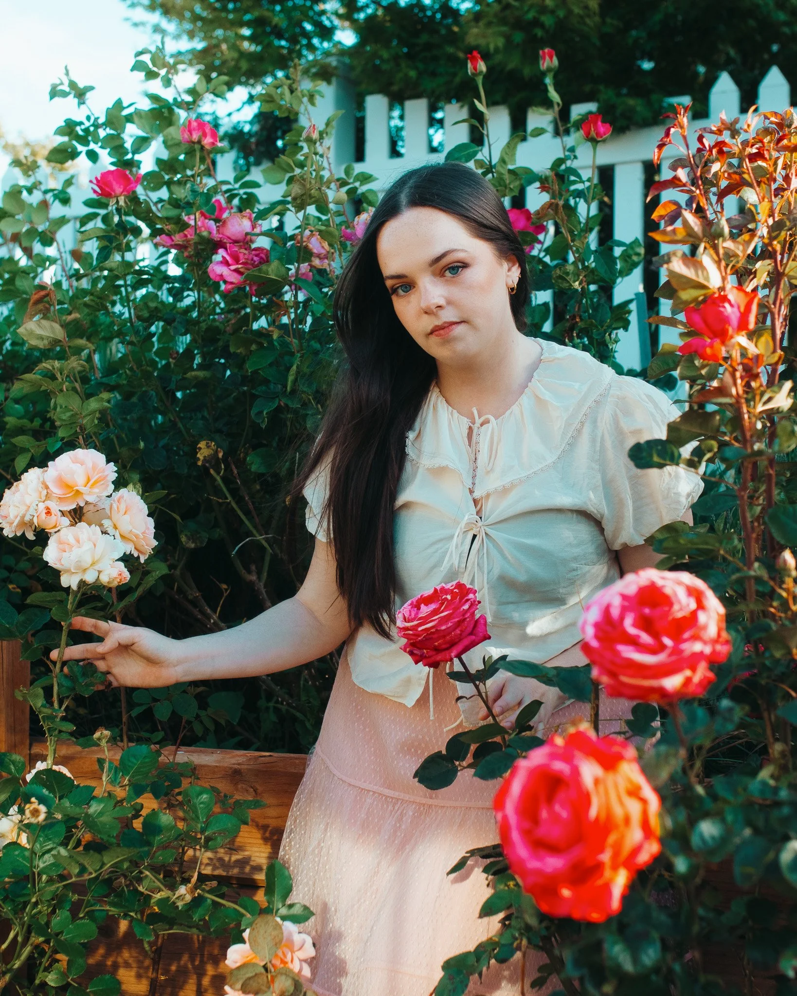 A woman with long dark hair and fair skin surrounded by blooming pink, red, and peach roses in a garden. She wears a cream-colored blouse with ruffled details and a light pink skirt. Behind her is a white wooden fence and green foliage.