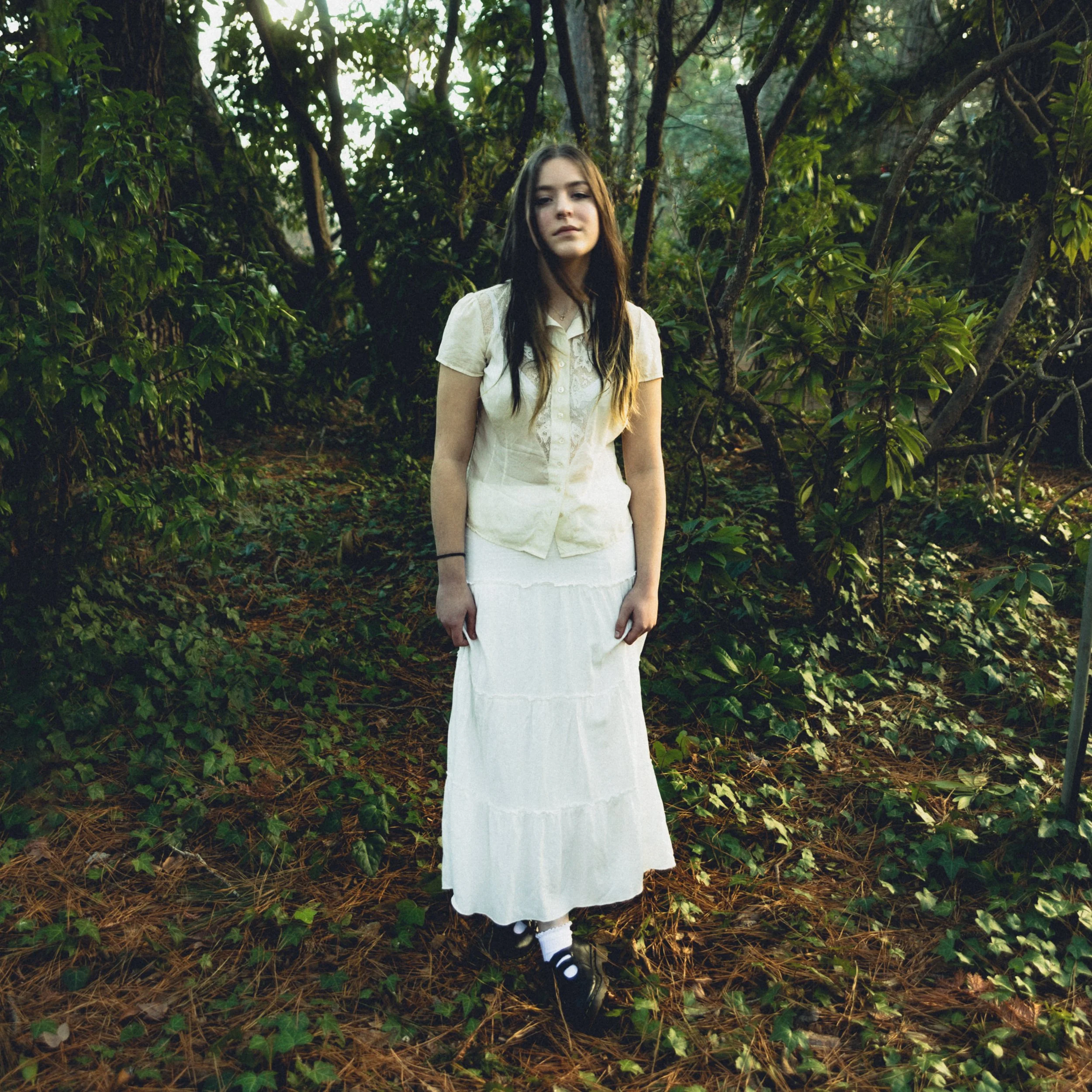A young woman with long brown hair wearing a white vintage-style dress, striped socks, and black shoes standing outdoors in a wooded area with green leaves and tree branches.