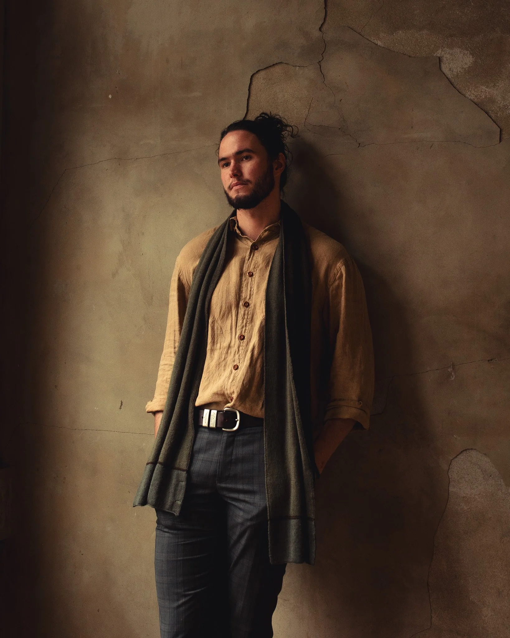 A young man with a beard and long hair in a bun leaning against a cracked and worn beige wall. He is wearing a tan button-up shirt, dark pants, and has a dark scarf draped over his shoulders.