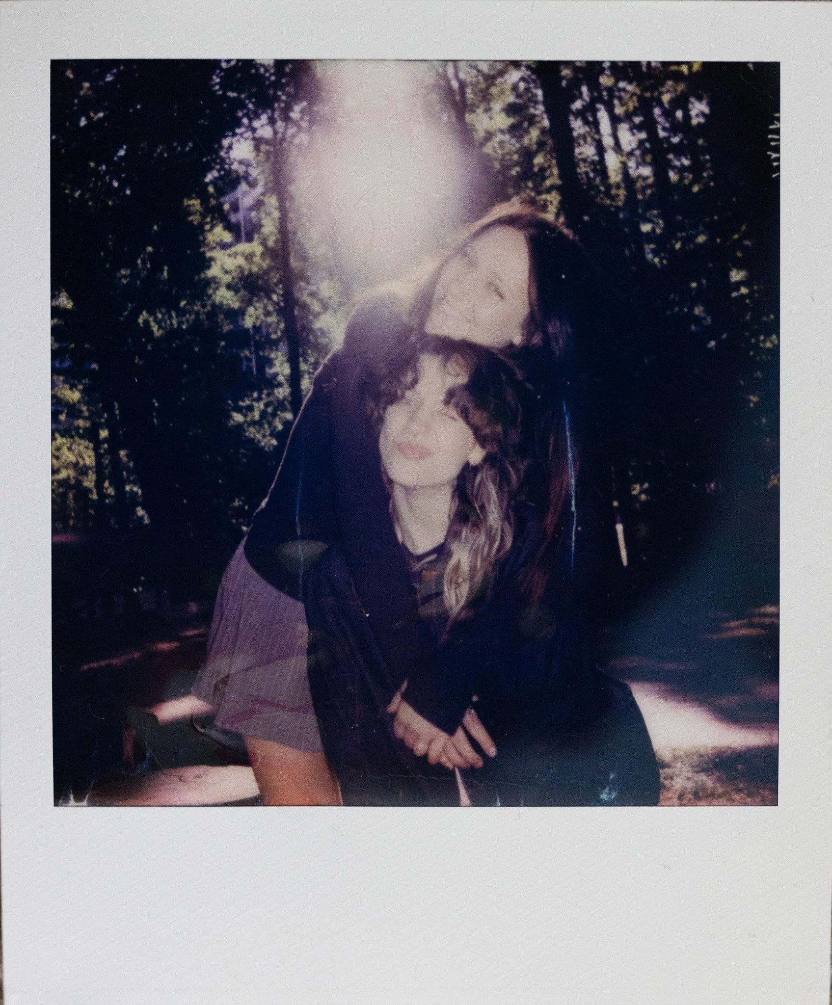A woman with long, dark, wavy hair and a woman with curly hair with glasses. They are outdoors with trees and sunlight in the background.