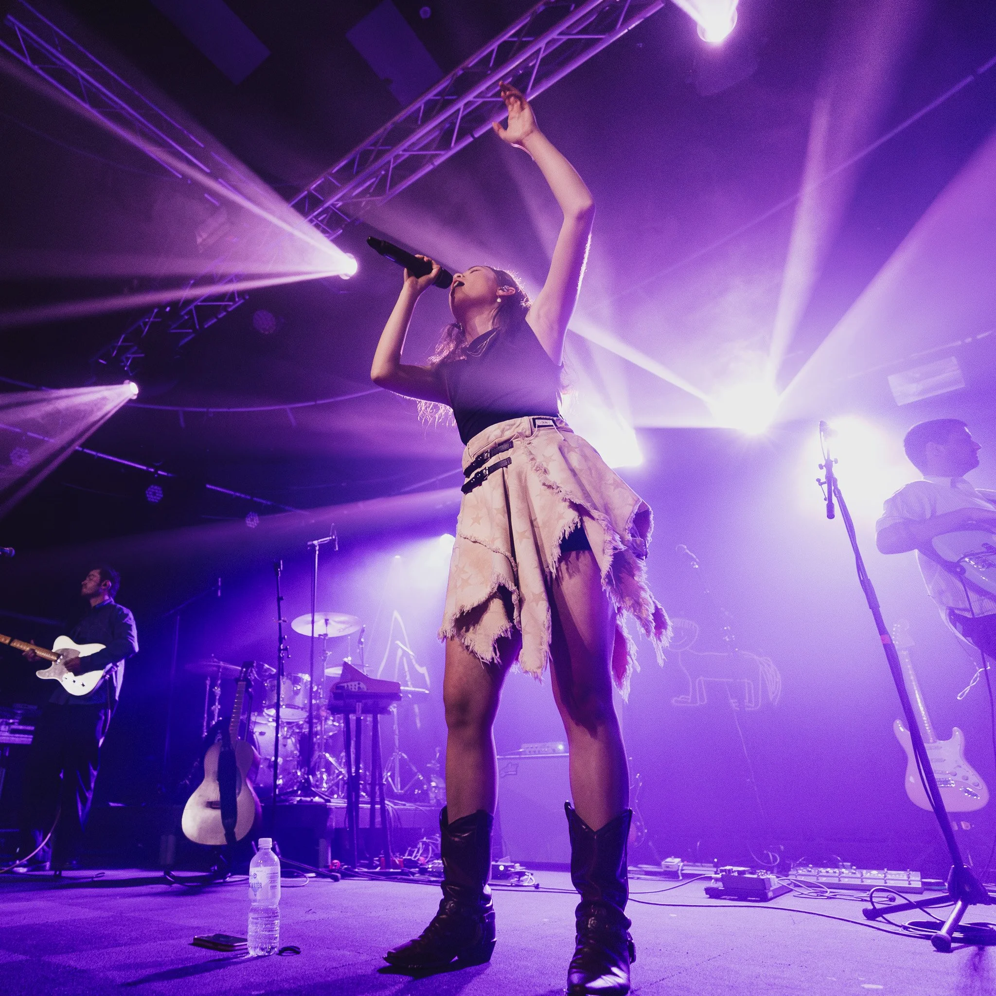 A female singer in a black top and beige skirt performs on stage with purple lighting, while a guitarist and other band members play behind her.