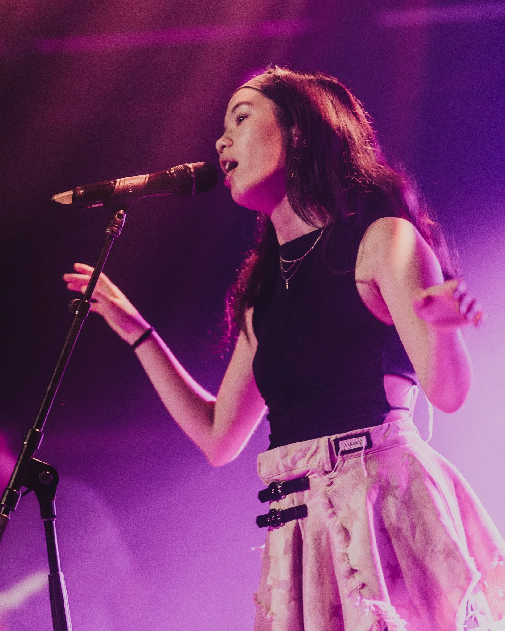 A woman with long dark hair singing into a microphone on stage, wearing a black top and light-colored ripped jeans, illuminated by purple stage lighting.