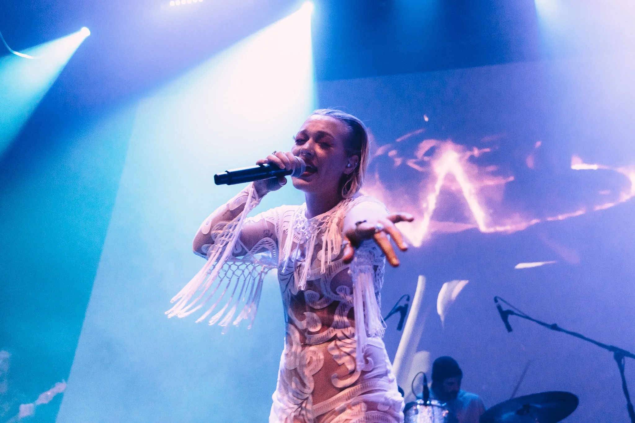 A female singer performing on stage in front of colorful, dramatic lighting, wearing a white outfit with fringes and reaching out towards the camera.