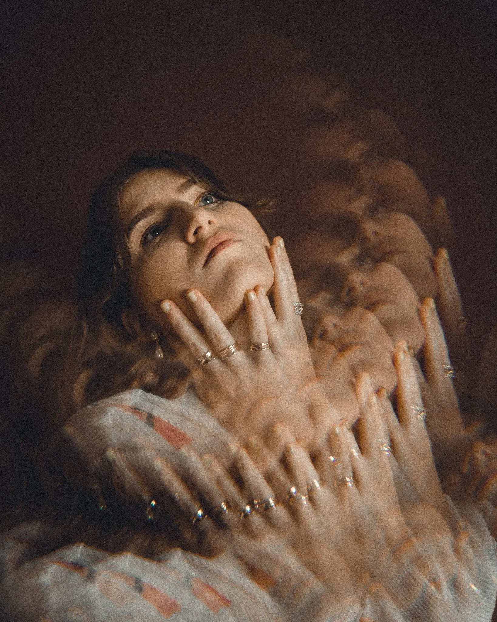 A woman with brown hair and blue eyes looking upwards, touching her face with her left hand, wearing rings, with a reflection of herself visible on a shiny, metallic surface.