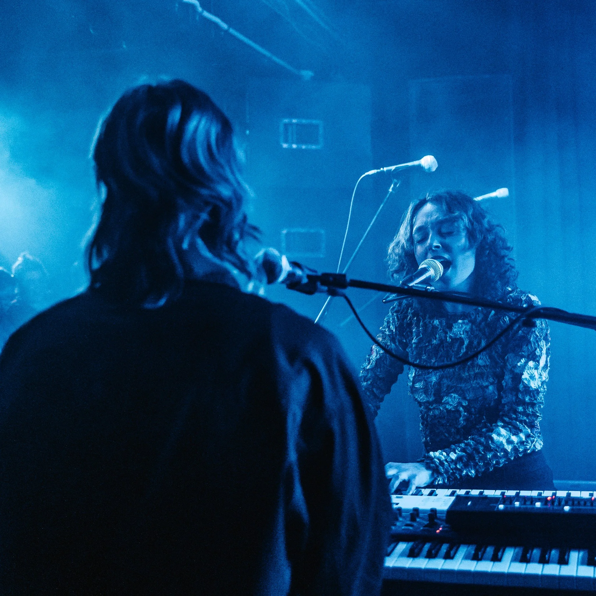 A close-up of a person with long brown hair sitting at a keyboard on stage, with a blue background and stage lighting. A person with curly hair singing into a microphone is visible in the background.