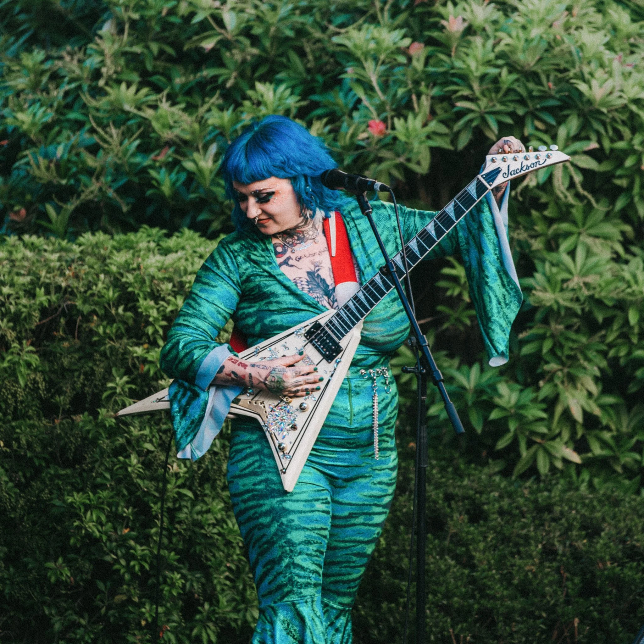 Person with bright blue hair playing an electric guitar outdoors in front of green bushes.