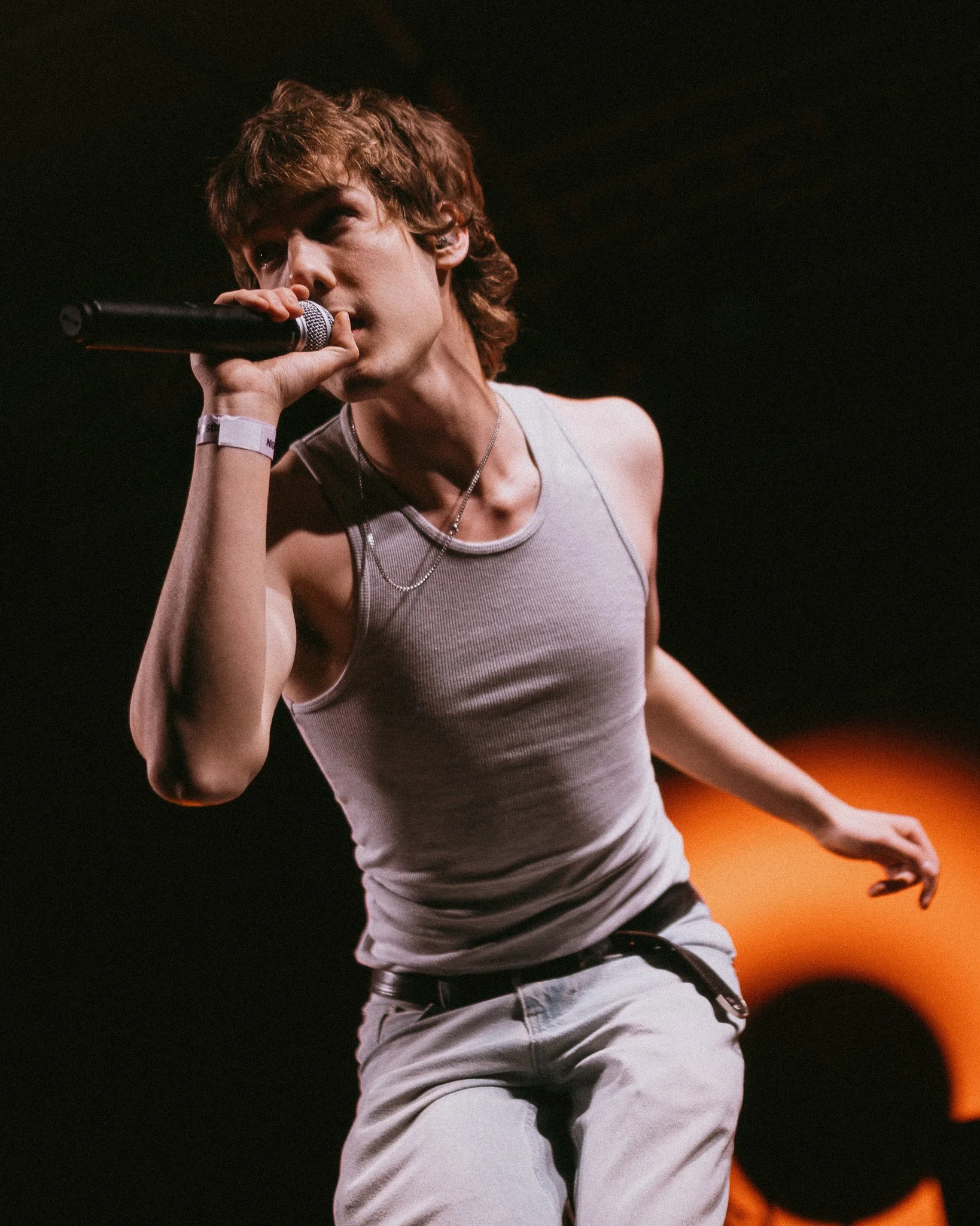 A young man with wavy, brown hair holds a microphone close to his mouth, performing on a dark stage with warm colored lighting.