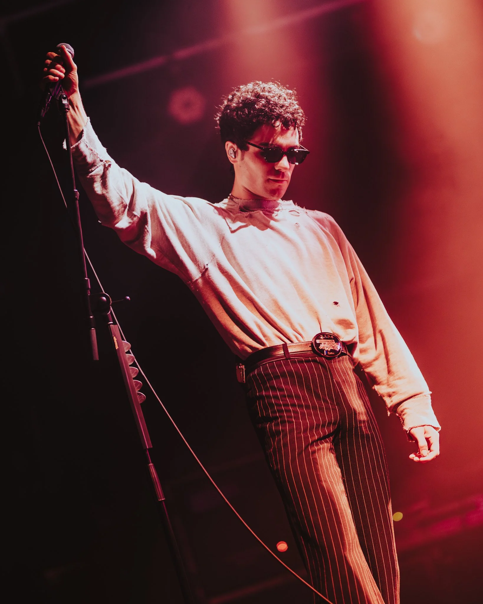 A male performer wearing sunglasses, a beige sweatshirt, and pinstripe pants holding a microphone on stage with pink and orange lighting.