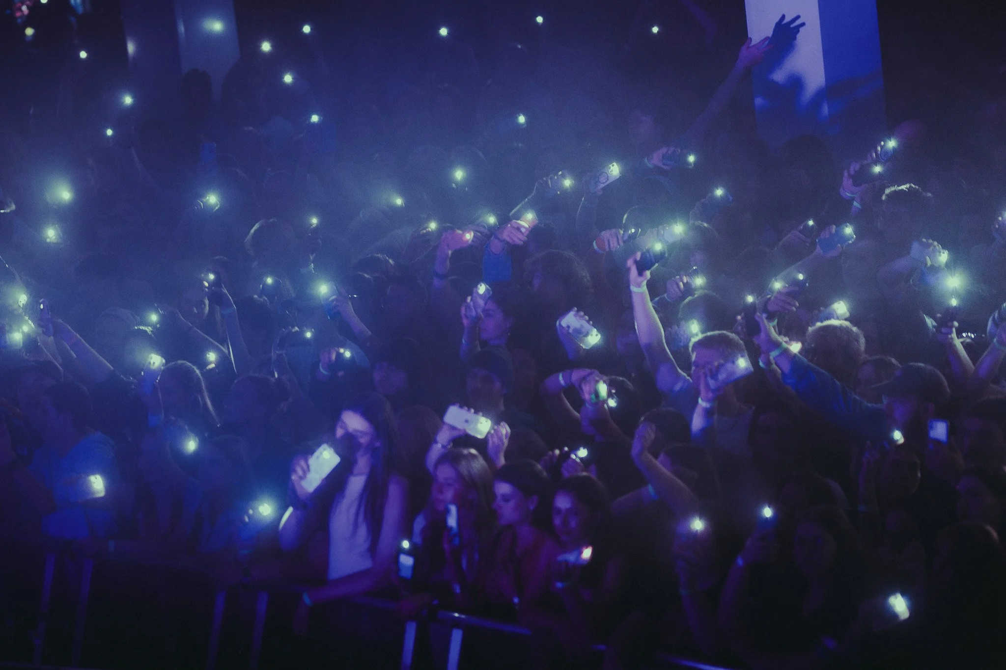 Crowd of people at a concert or event holding up their phones with flashlight lights on, in a dark room with blue lighting.