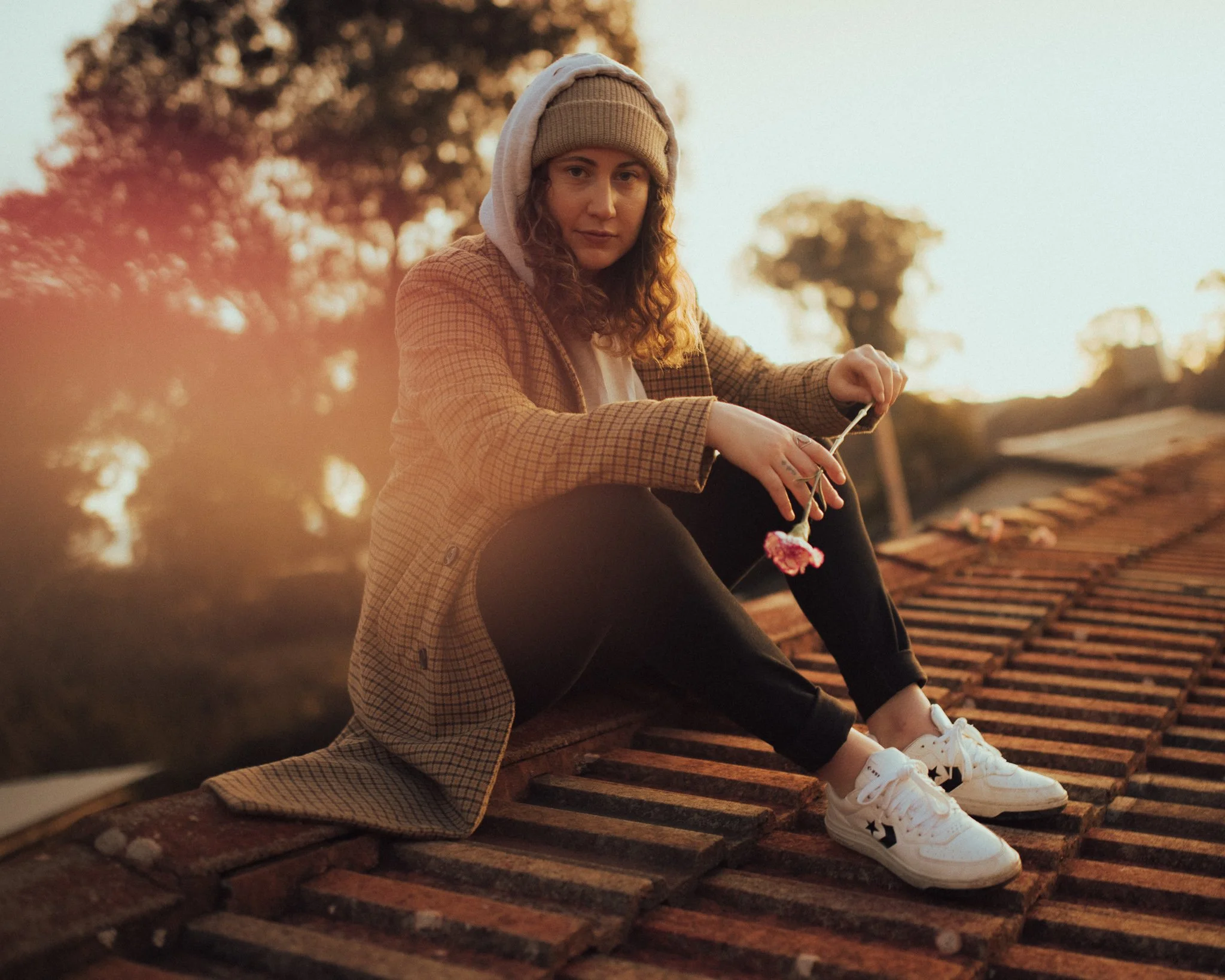 A young woman with curly hair sitting on a wooden railway track during sunset, wearing a beige knit hat, hoodie, checked coat, black leggings, and white sneakers with black star patterns.