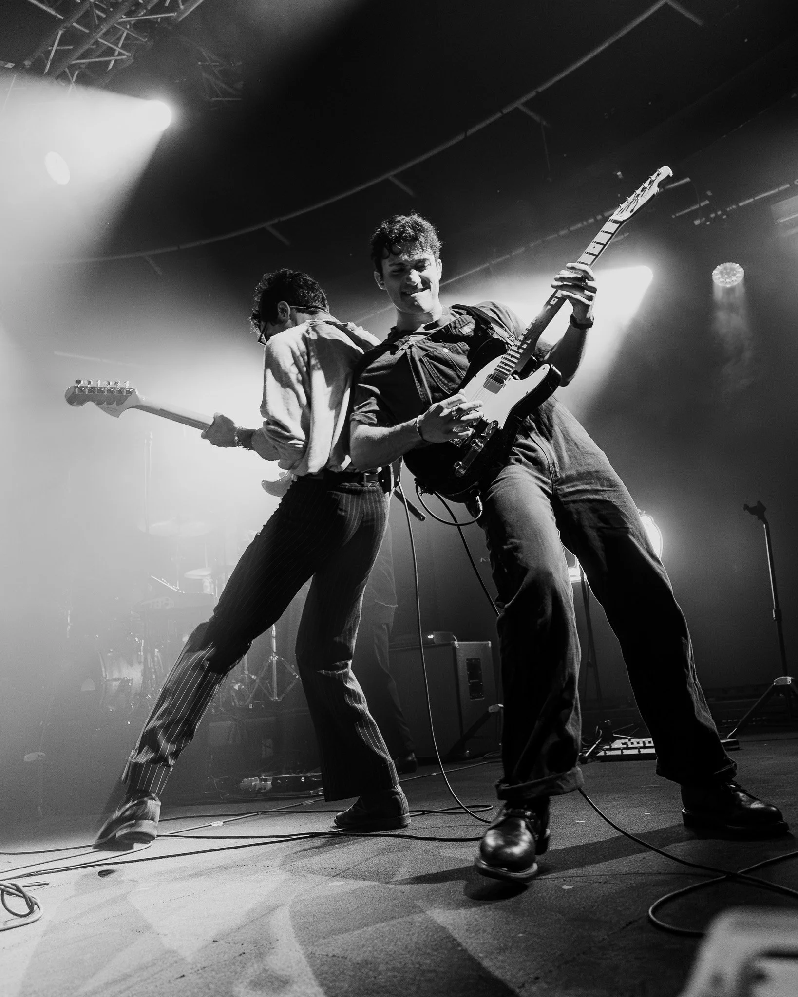 Black and white photo of two musicians on stage playing electric guitars, with stage lights shining down on them.