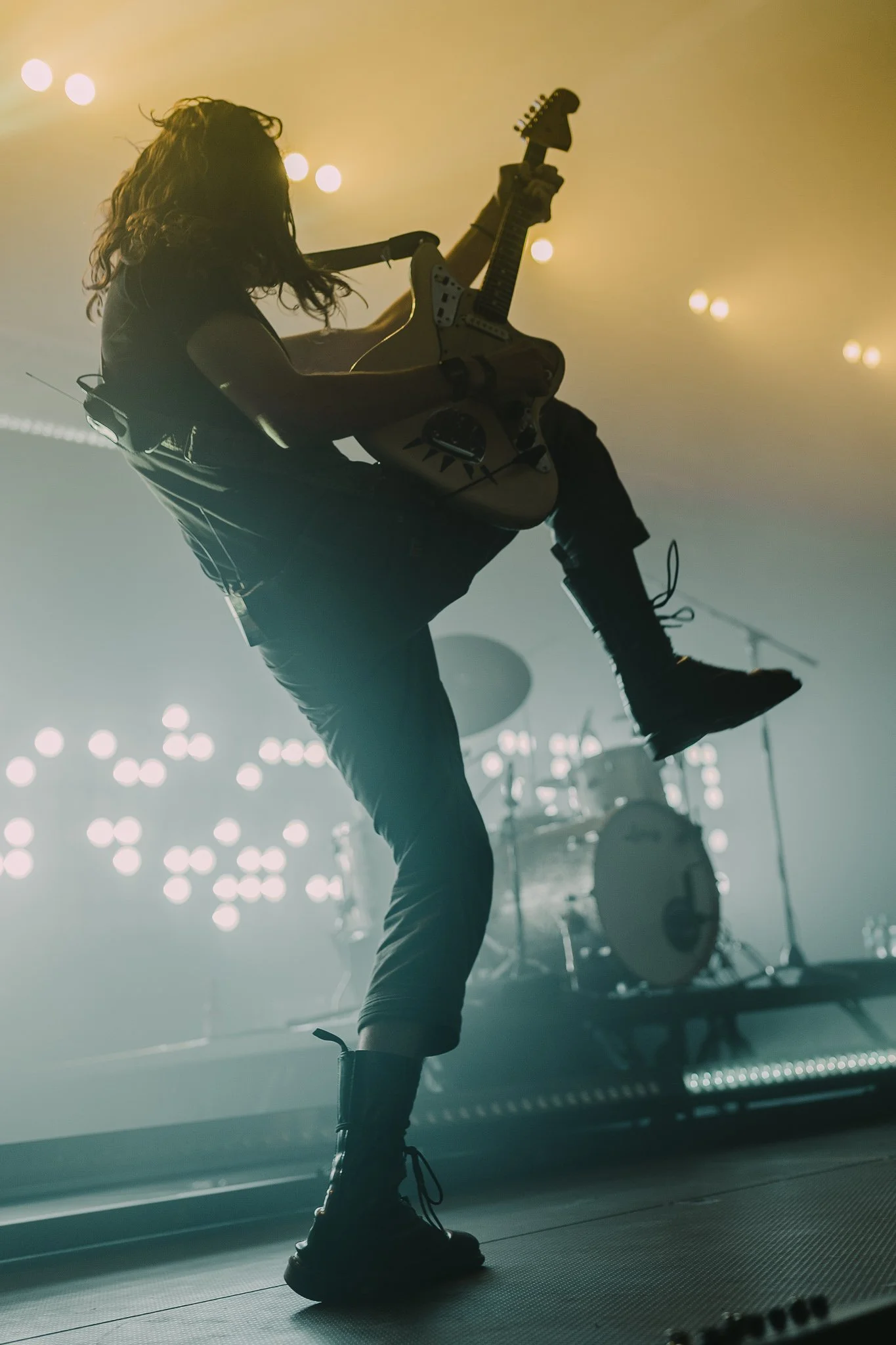 Silhouette of a guitarist playing on stage with bright stage lights and a drum set in the background.