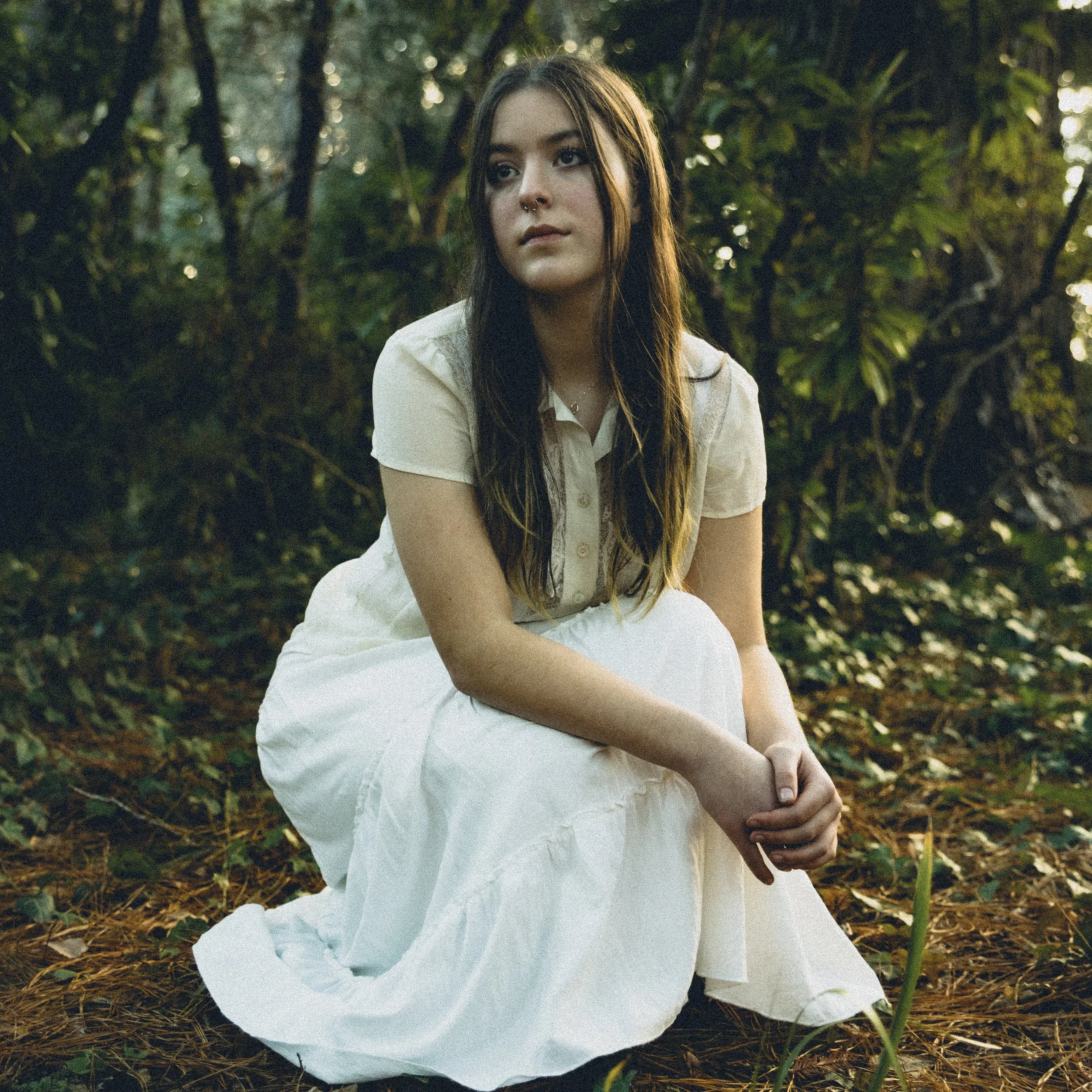 A young woman with long brown hair and a septum piercing sitting on the ground in a forest surrounded by greenery and trees.