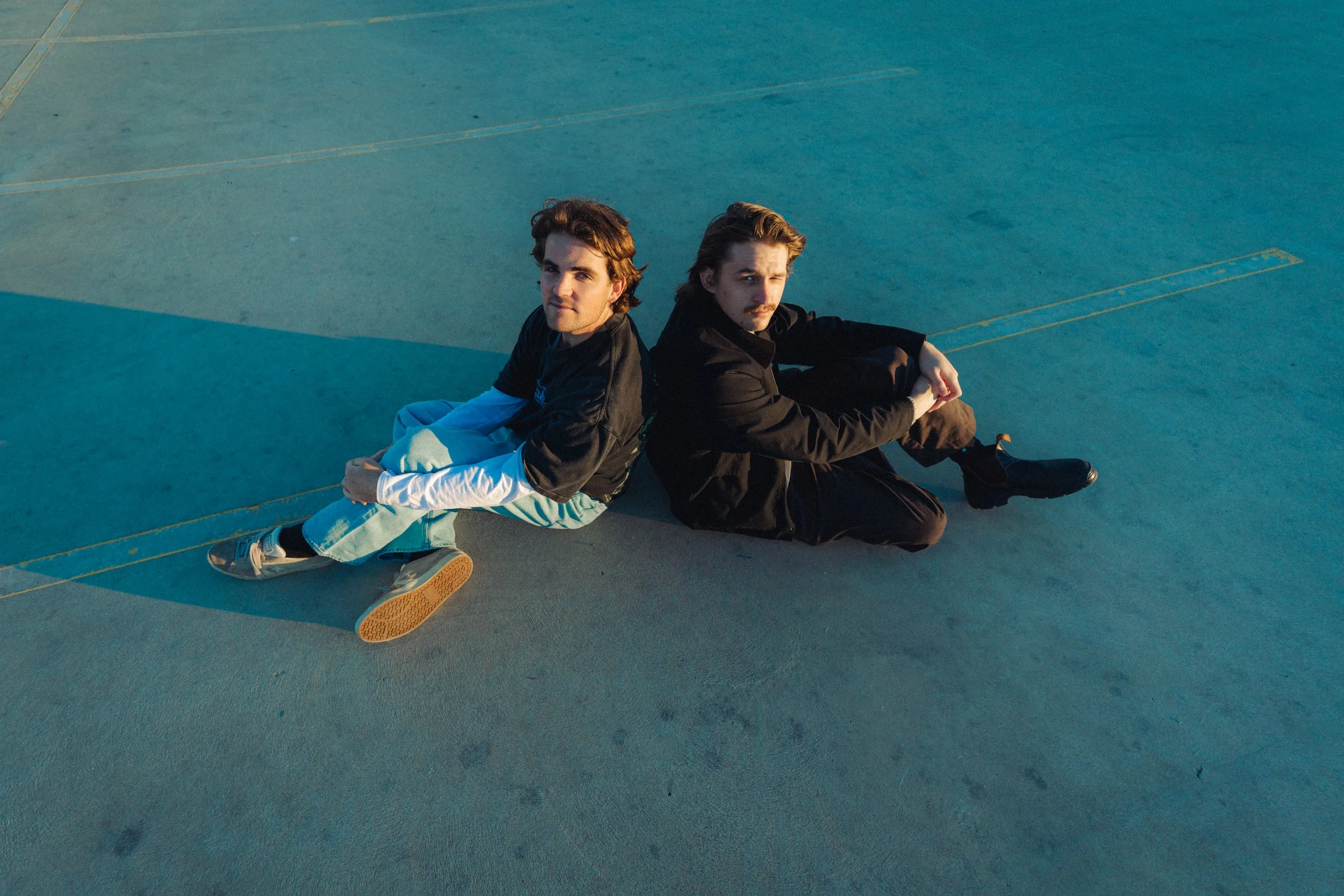 Two young men with brown hair sitting on a concrete surface, facing opposite directions and looking at the camera, with a yellow line in the background.