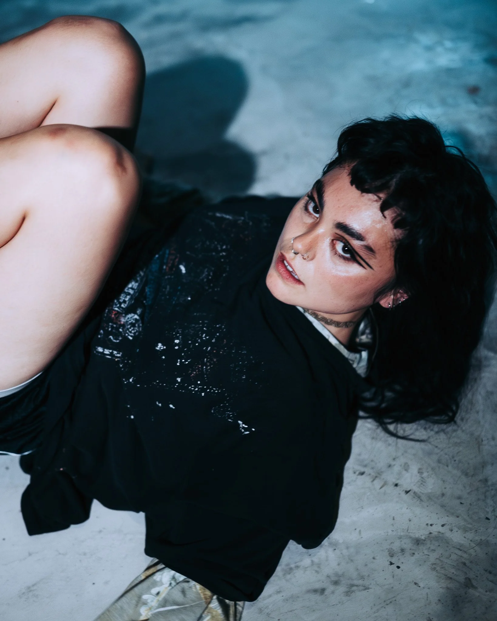 A person with dark, tousled hair lying on the ground, wearing black clothing with a shiny texture. They have piercings on their nose and ears, and are looking up at the camera with a neutral expression. The background is a gravel or dirt surface.