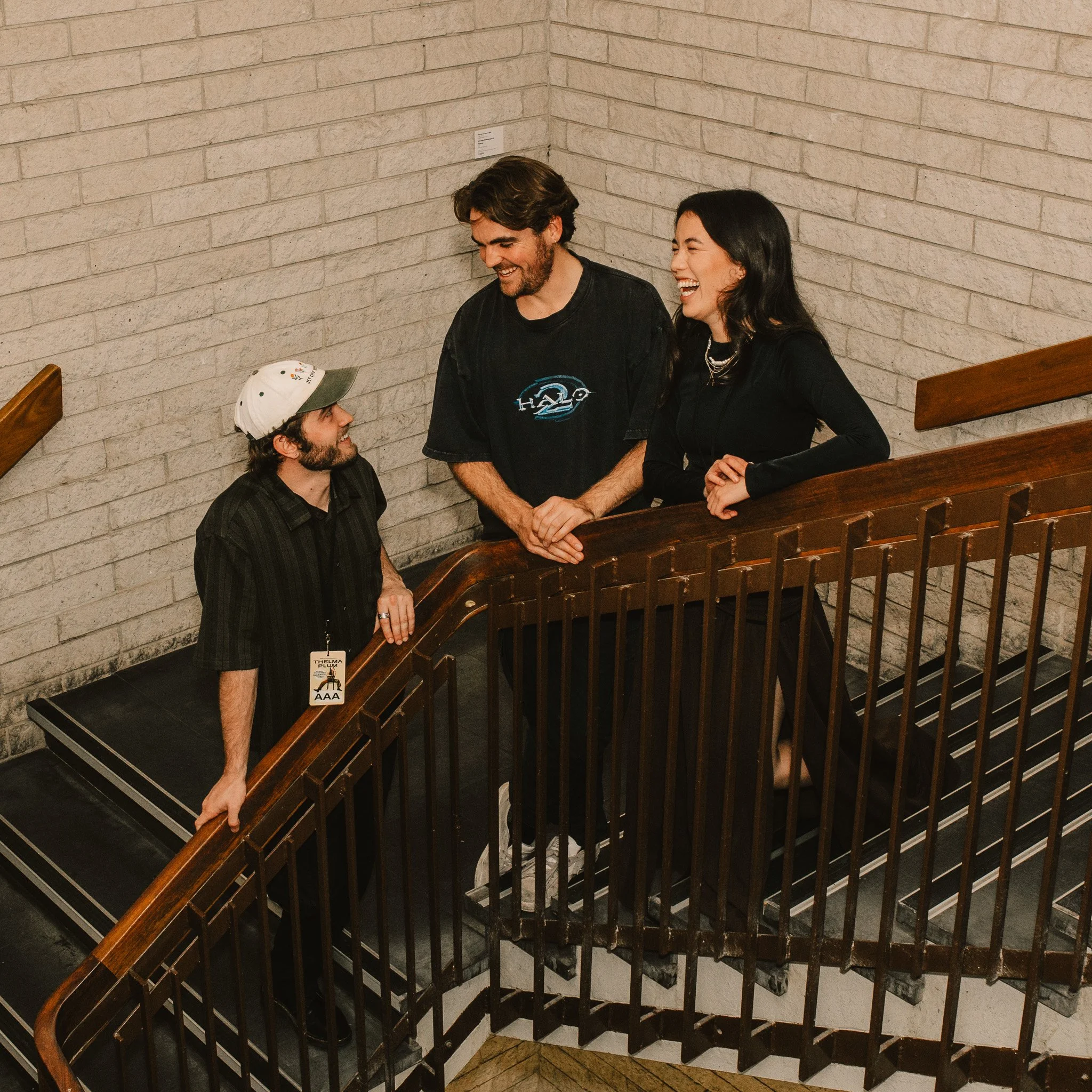 Two men and a woman smiling and talking near a staircase with a brick wall in the background.
