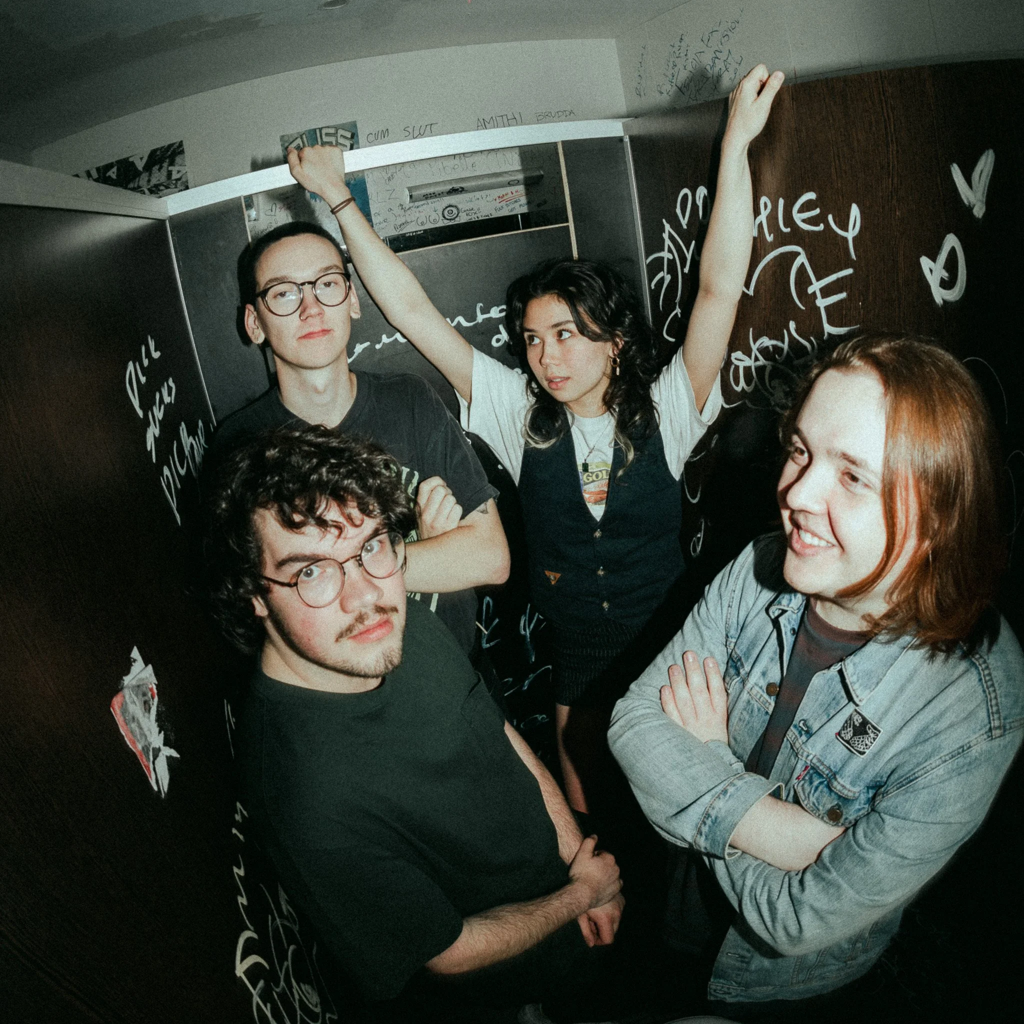 A group of young people standing inside a room with black walls covered in white handwritten text and graffiti. Two of them, a man wearing glasses and a woman with dark hair, are visible. The man has his arms crossed and looks directly at the camera,