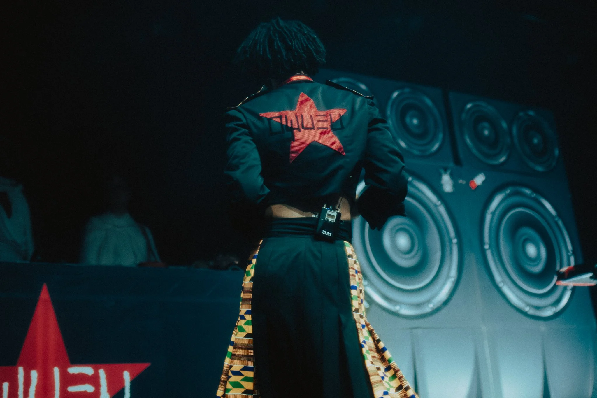 A person with curly hair is seen from behind, wearing a black jacket with a red star and the word 'LOYALTY' on the back, standing in front of a wall of large speakers at night.