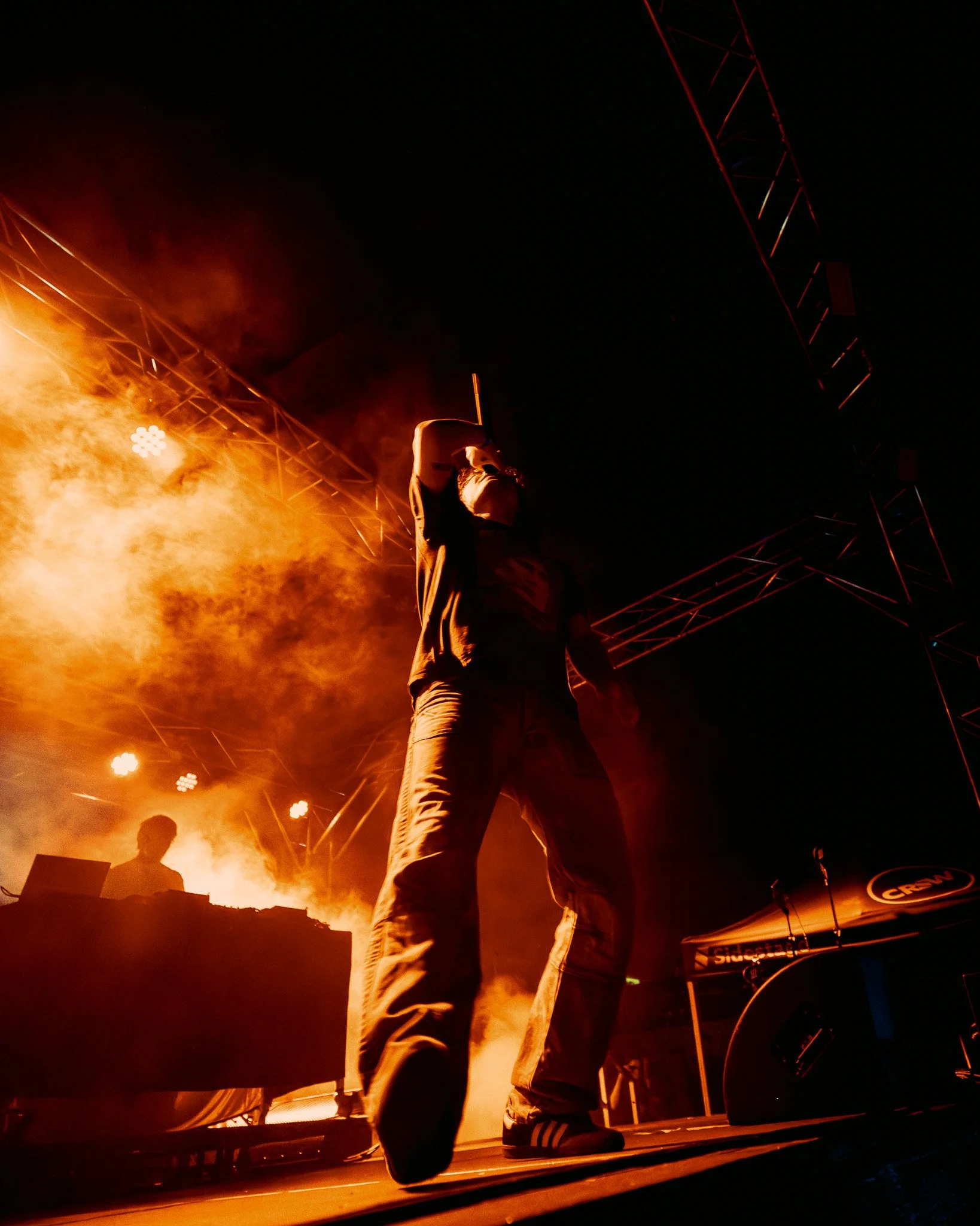 A performer on stage with a microphone, backlit by bright orange flames, surrounded by stage equipment and red lighting trusses.