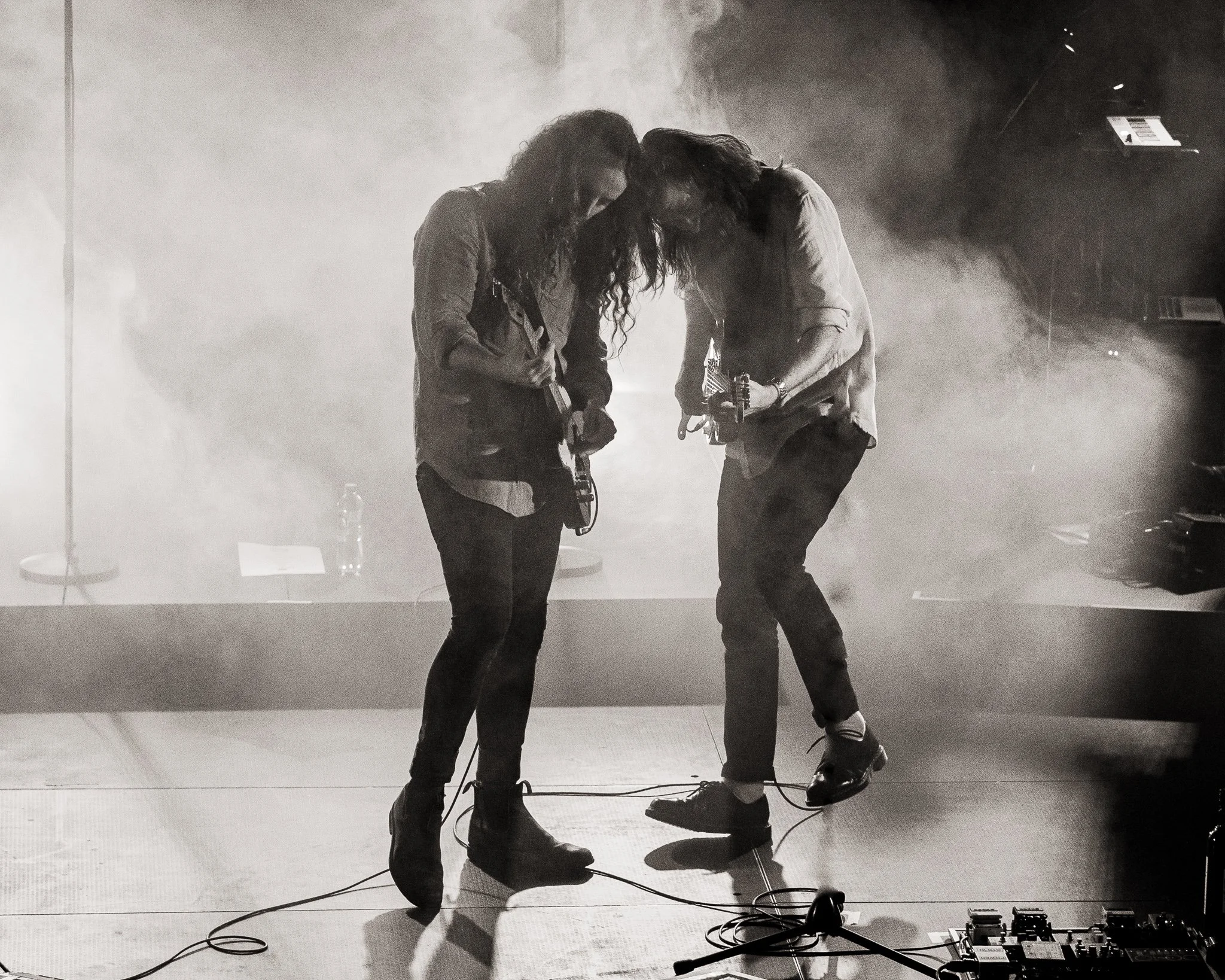 Two musicians with long hair huddled together, appearing to be sharing a moment during a performance, surrounded by stage equipment and fog.