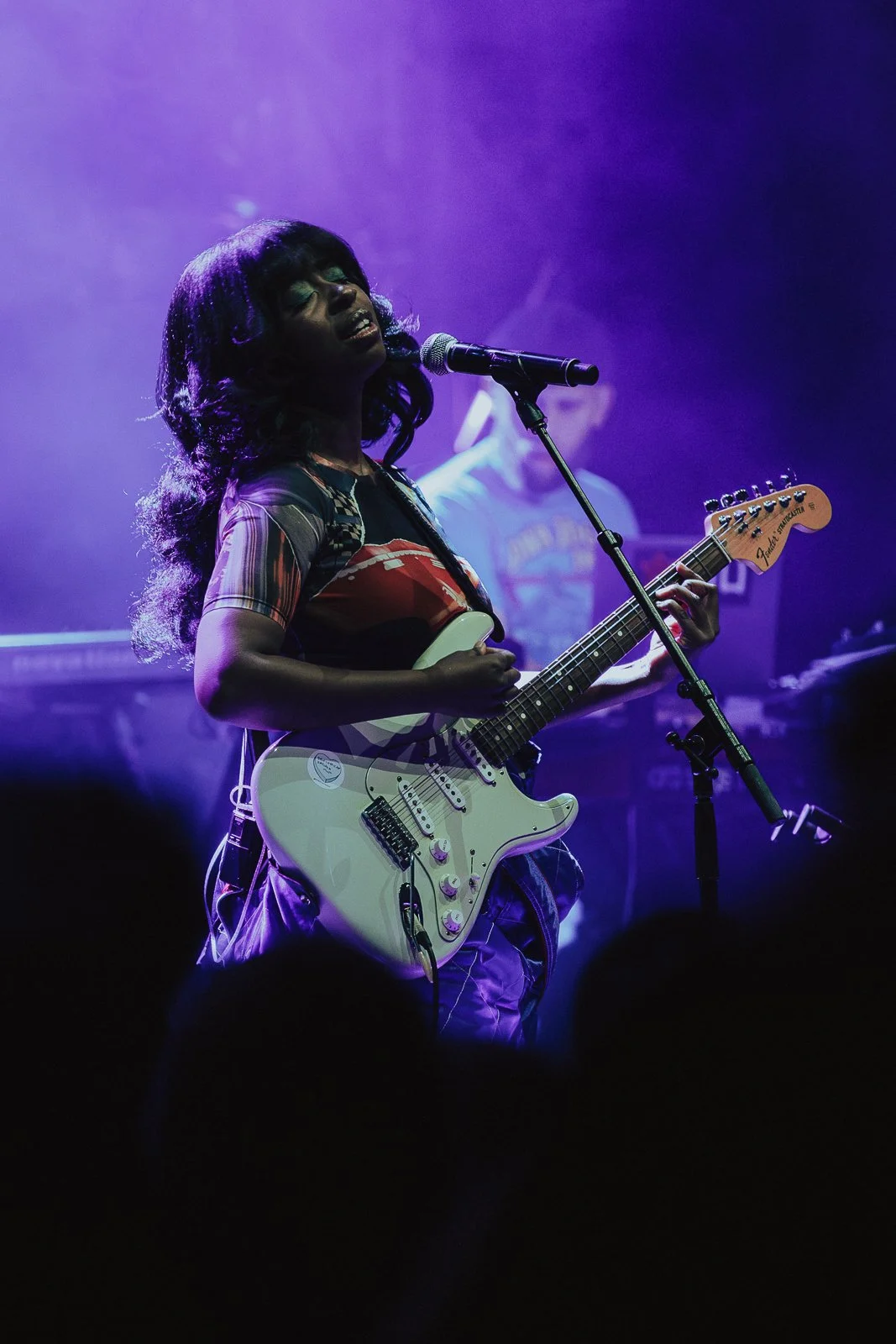 A female musician singing and playing an electric guitar on stage with purple lighting, with a male musician in the background playing a keyboard.