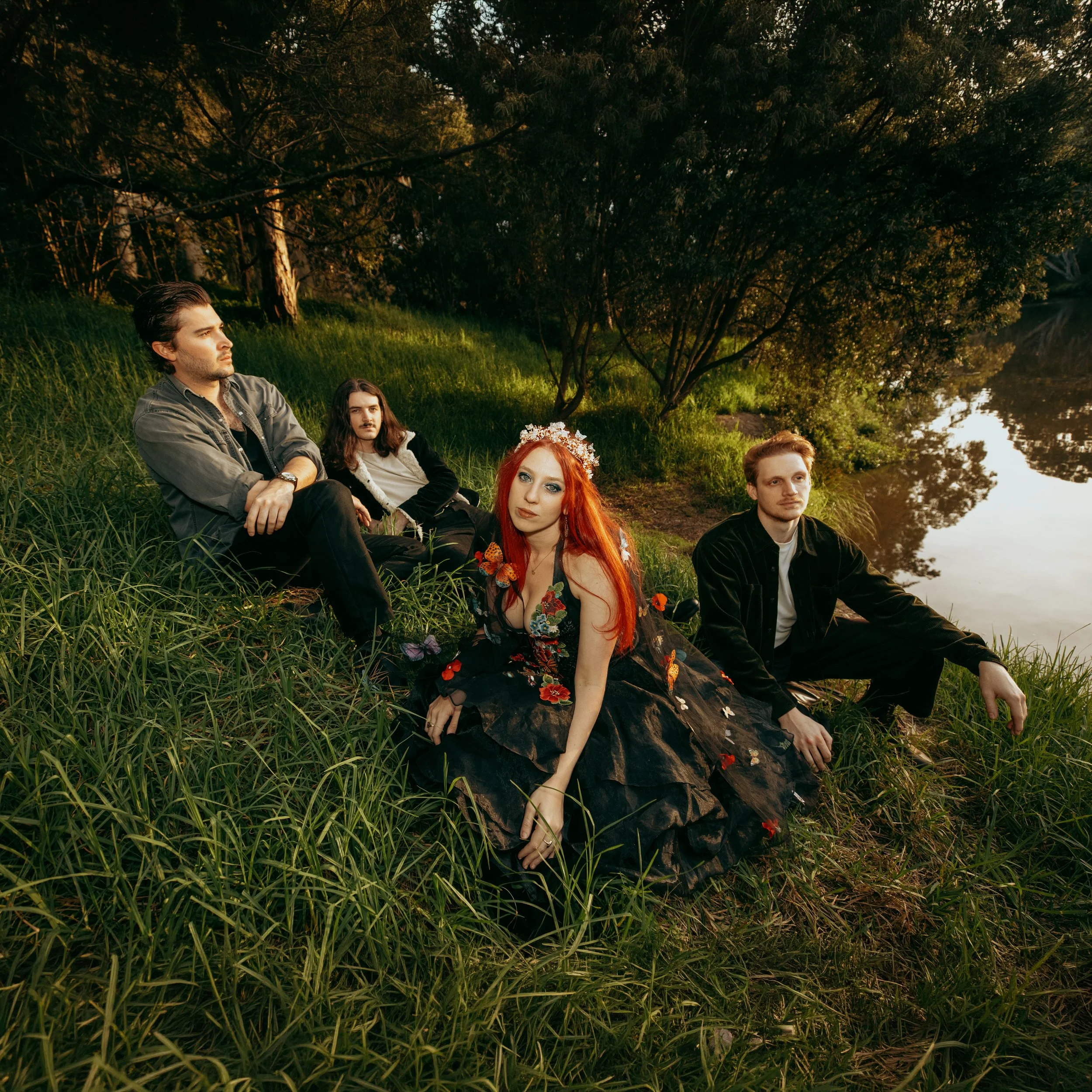 Group of five people sitting on grass near a river under trees in a park during sunset, with one woman wearing a floral headpiece and dress, others dressed casually.