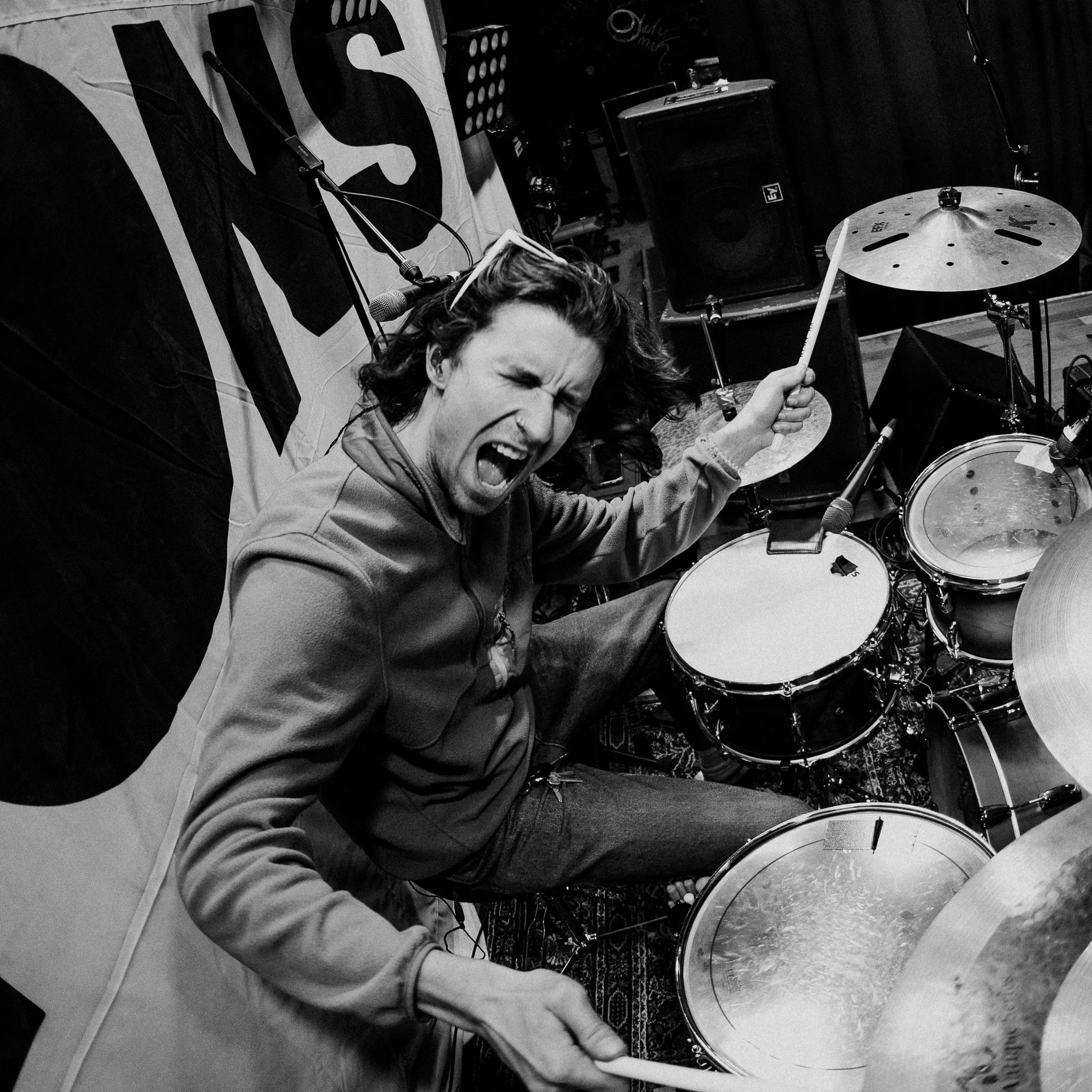 A woman passionately singing into a microphone with her eyes closed, holding a drumstick. She is surrounded by musical equipment including drums, an amplifier, and microphones. The image is in black and white.