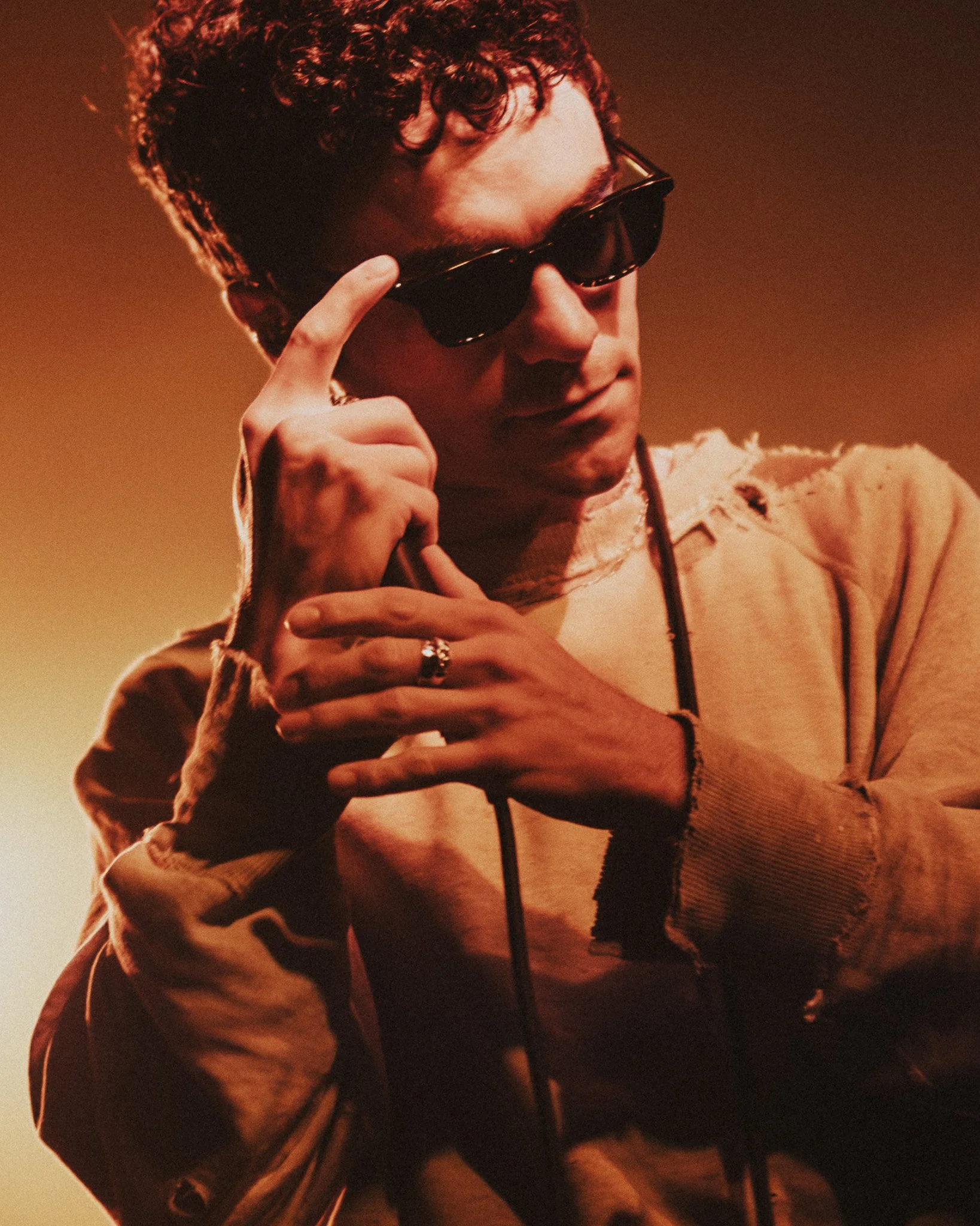 A person with curly hair wearing sunglasses, a beige jacket, and a ring on their finger. They are holding their glasses with their left hand and touching their chest with their right hand. The background is warm-toned and slightly blurred.