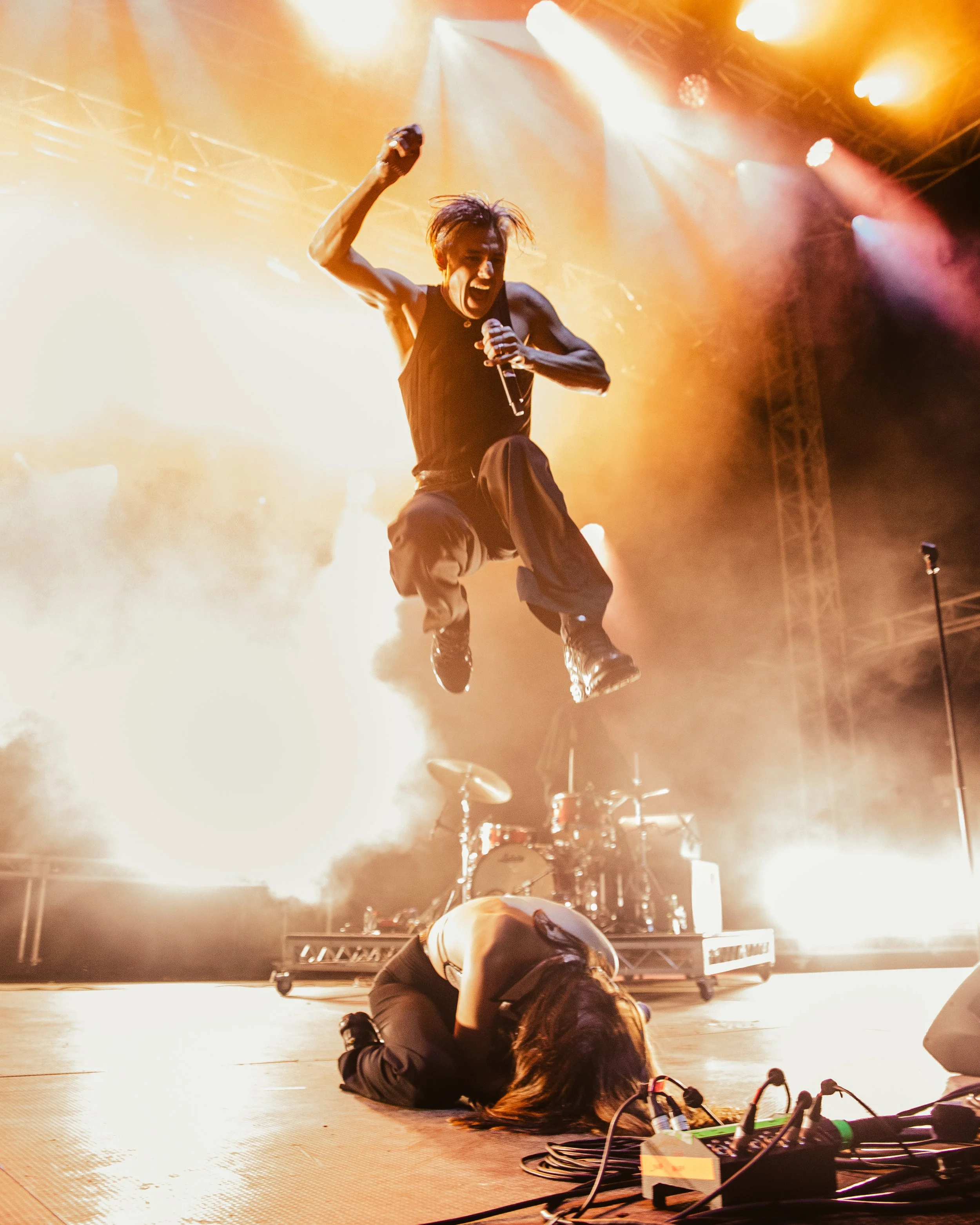 A performer, possibly a singer, jumping in the air on stage with bright, colorful lights and smoke in the background. An audience member or crew is visible in the foreground on the floor, and musical equipment is nearby.