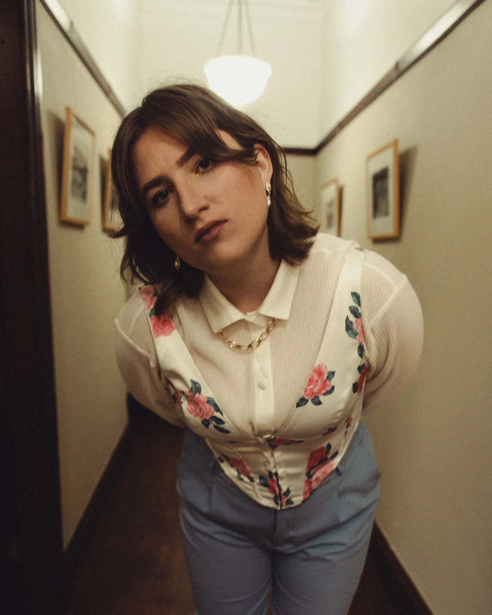 A woman with shoulder-length brown hair, wearing earrings, a necklace, a cream-colored blouse with floral patterns, and blue pants standing in a hallway with framed pictures on the wall.