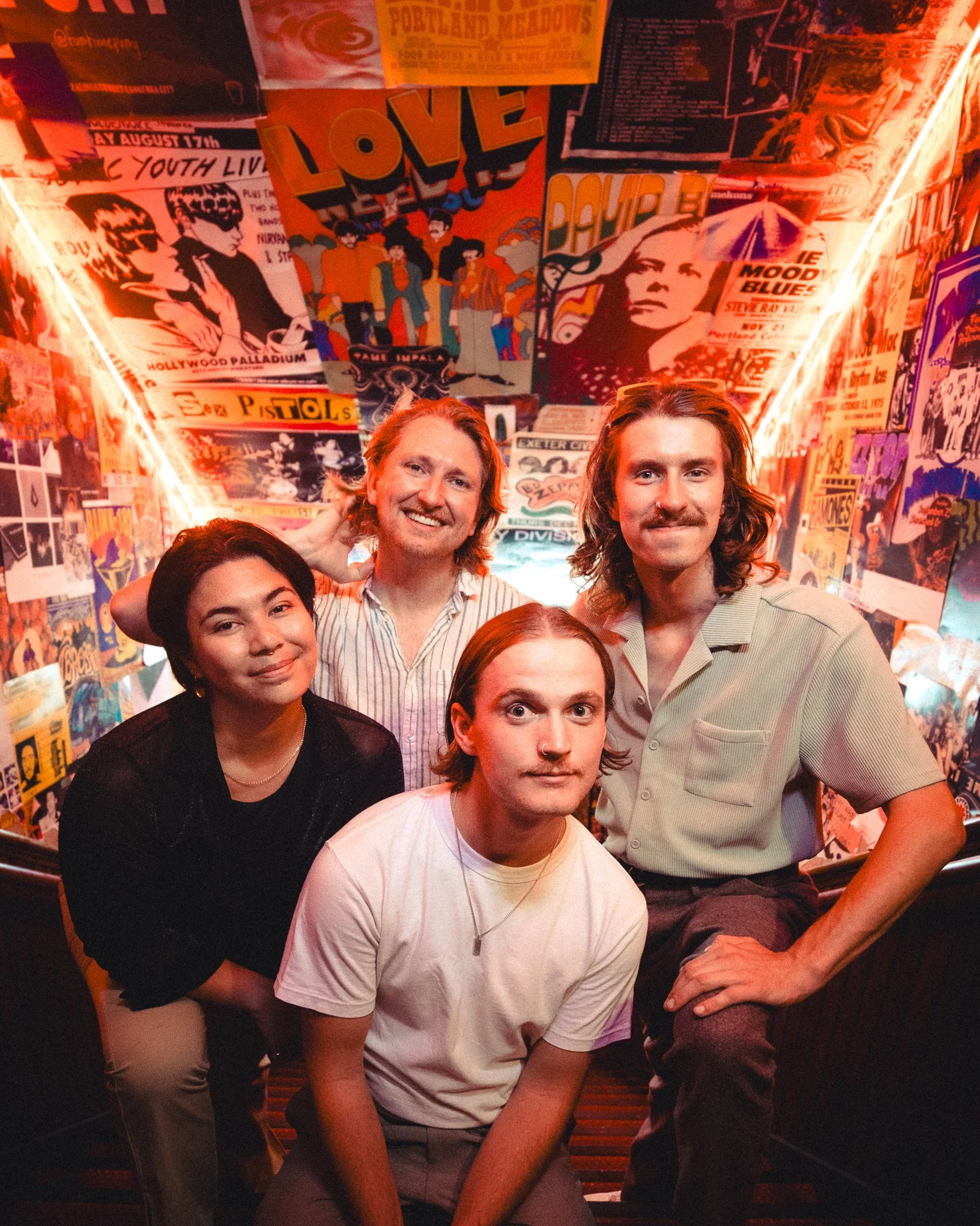 Group of five people posing together in front of a wall covered with colorful posters and images from music and pop culture, in a lively, vibrant setting.