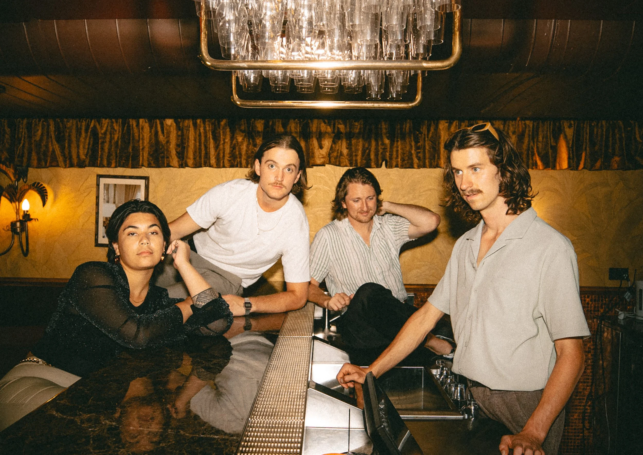 Four young adults with varied hair colors and styles, seated around a bar counter in a retro-style room with warm lighting, wood paneling, and decorative curtains.