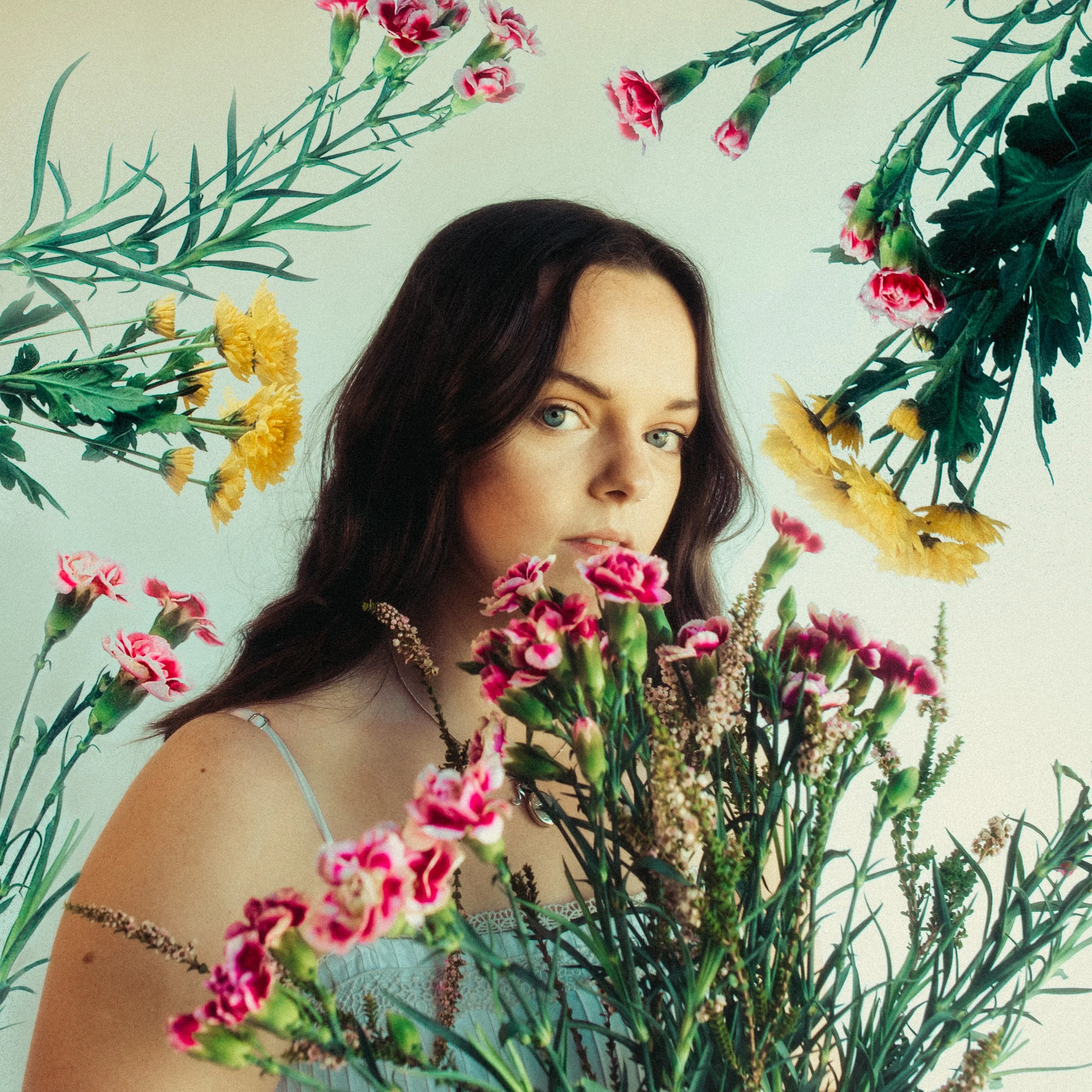 A woman with dark hair and blue eyes surrounded by colorful flowers, looking at the camera.