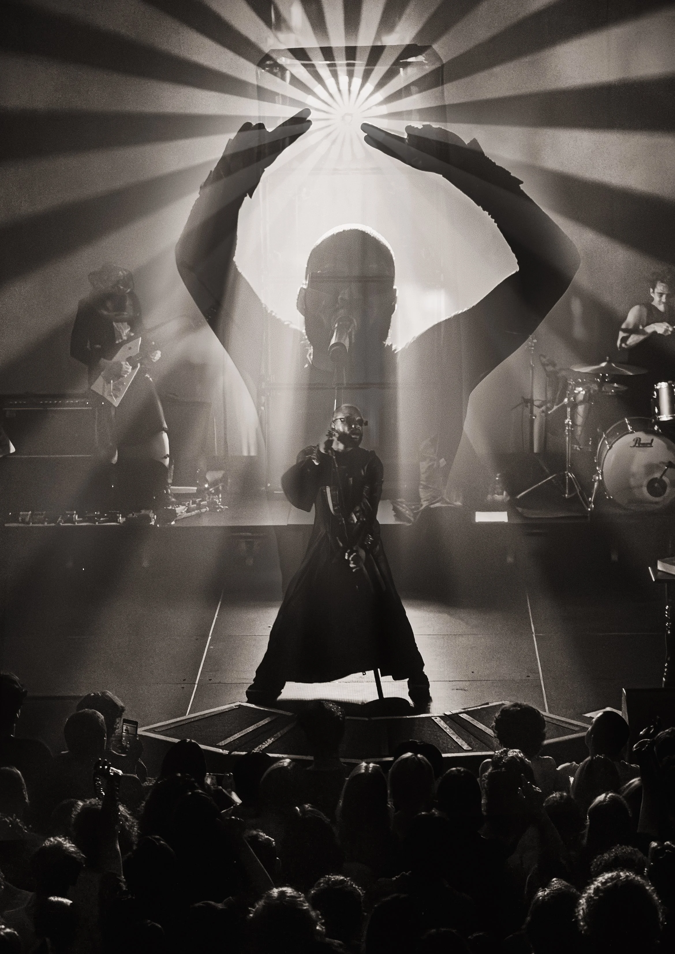 Black and white concert scene with a performer in front of the stage, raising arms with hands forming a triangle shape, behind a large screen showing a silhouette of a person with visual effects resembling rays of light, and musicians on the sides pl