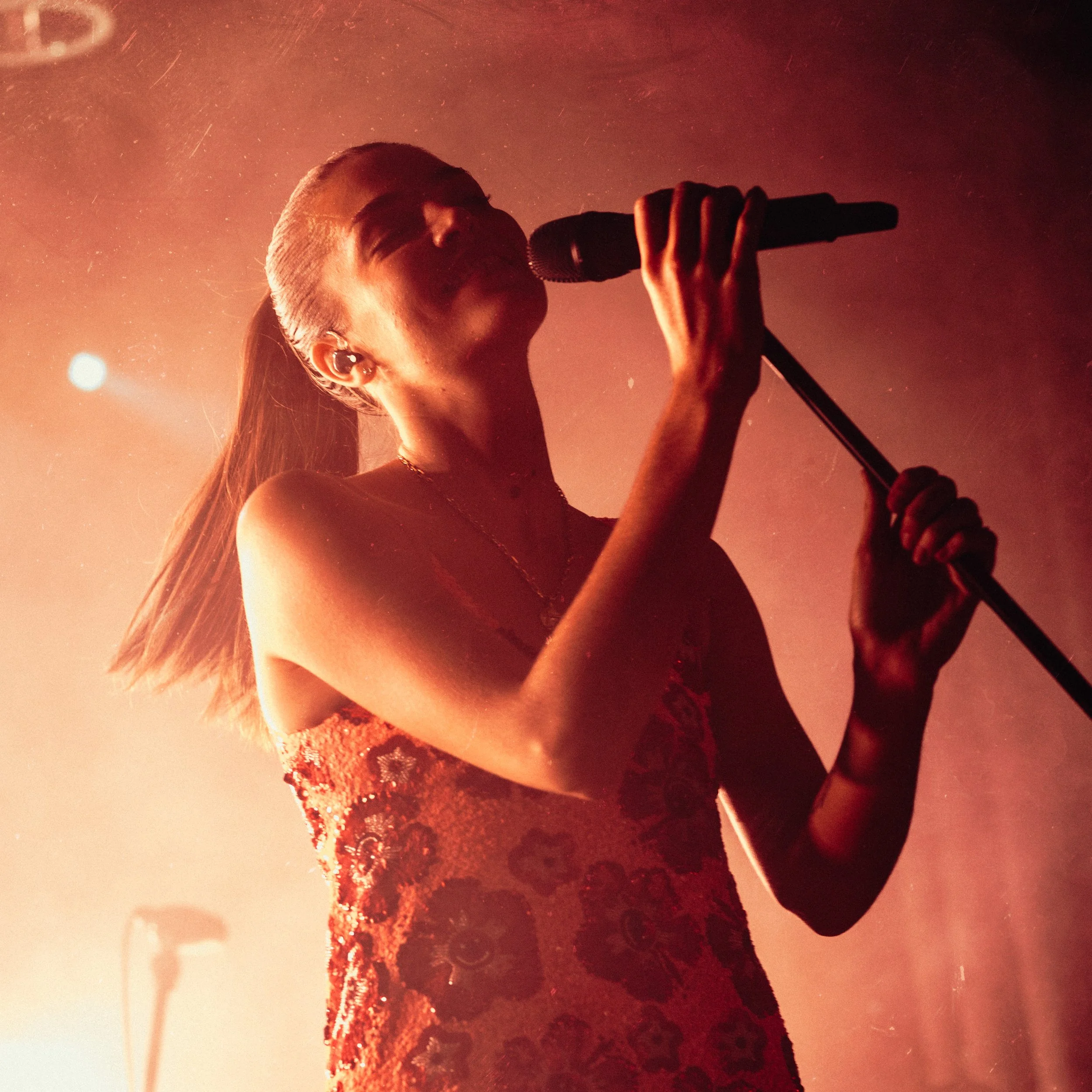 A person singing into a microphone, dressed in a lace outfit, with their hair tied back, against a red-orange background, captured from a low angle.