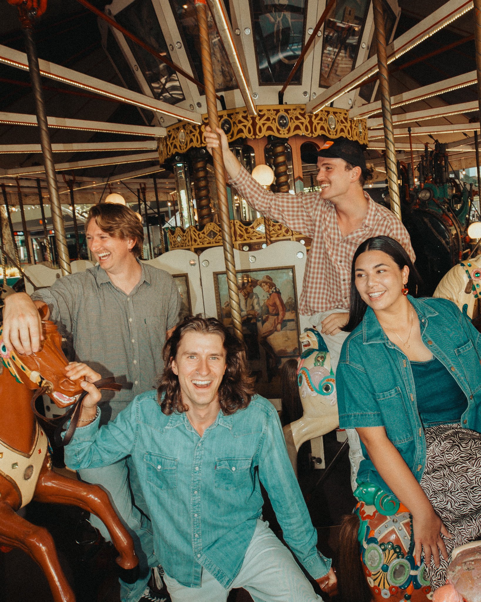 People smiling and enjoying a carousel ride at an amusement park or fair. The image features a man in a plaid shirt holding onto a carousel pole, a woman with earrings and a sleeveless denim jacket, and another person with long hair in the foreground
