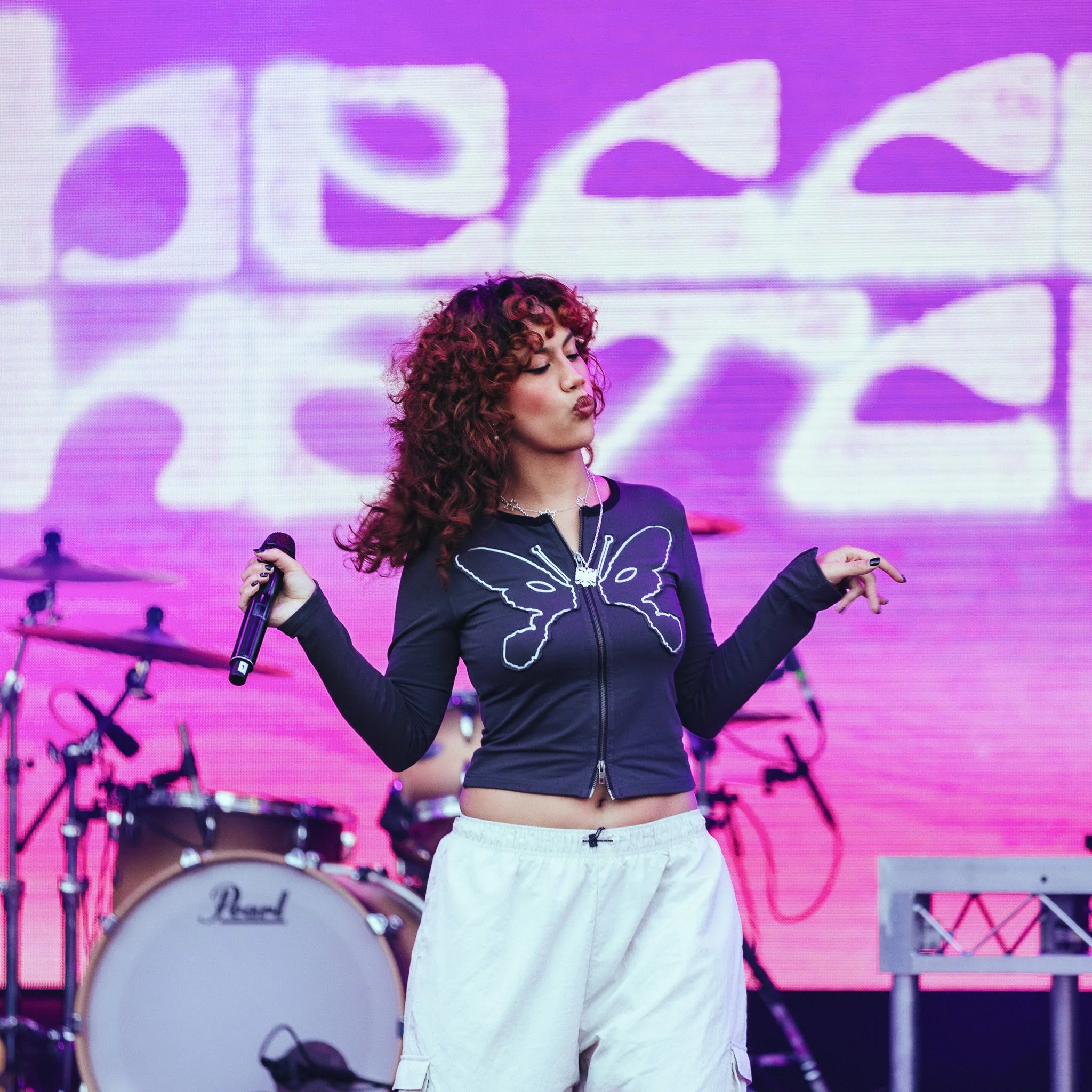 A young woman with curly hair wearing a black shirt with a butterfly design, white pants, and holding a microphone, standing on a stage with pink and purple lighting and musical instruments in the background.