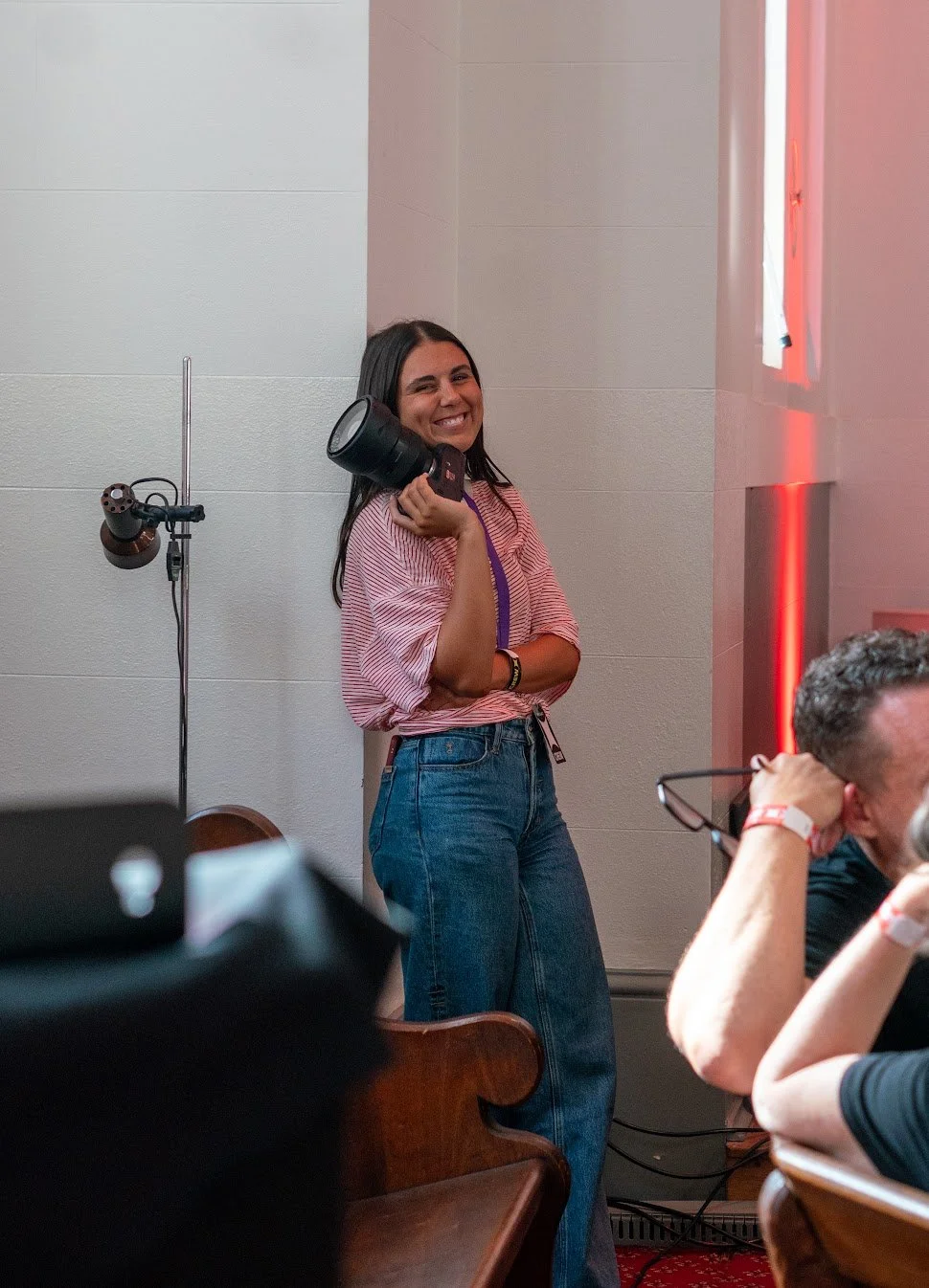 A smiling woman holding a camera in her right hand, standing in a room with light-colored walls and red-lit decor, while other people sit at a table nearby.